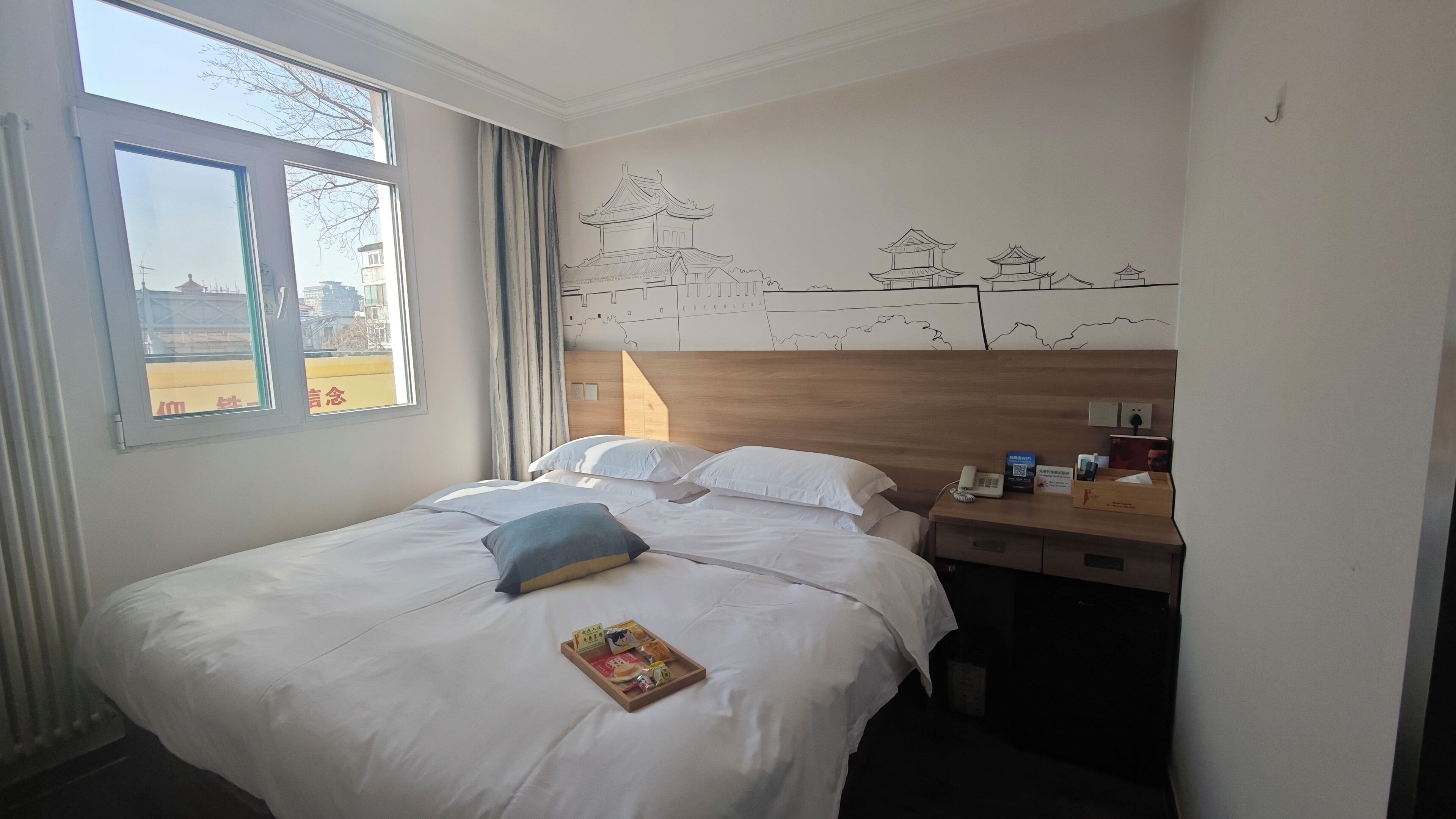 A hotel room with a large bed, a wooden headboard, a bedside table with a phone and toiletries, and a window with a view of buildings.