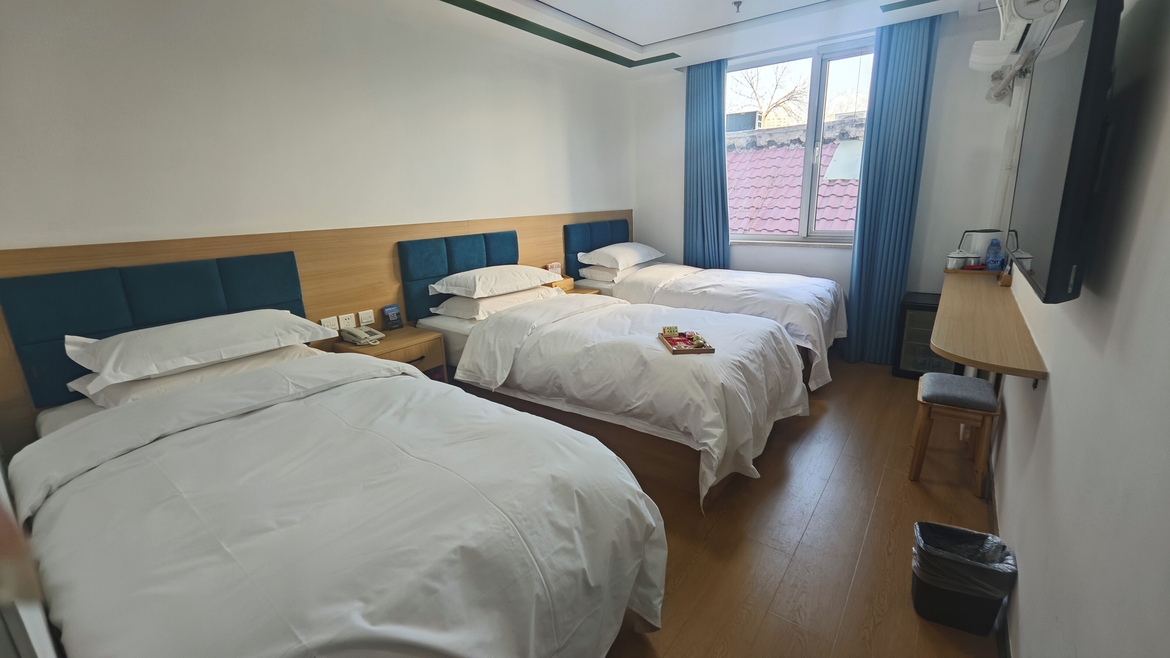 A hotel room with two beds, a wooden headboard, a small table, a trash bin, and a window with curtains.