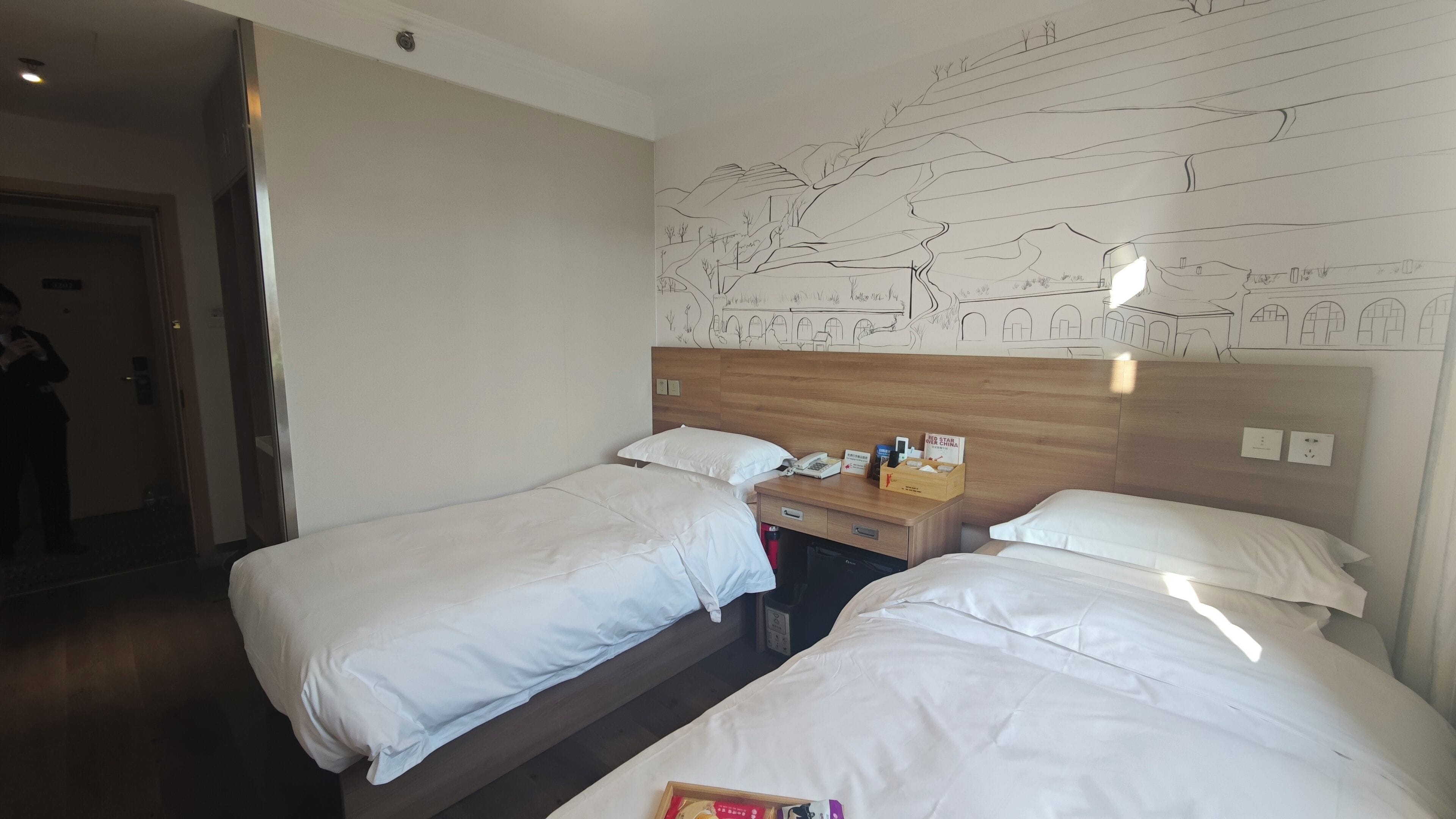 A hotel room with two beds, a wooden headboard, a nightstand with a phone and toiletries, and a wall mural.