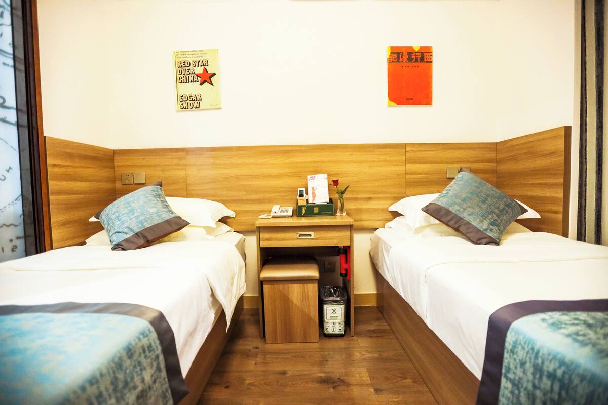Habitación de hotel con dos camas, un cabecero de madera, una mesita de noche, un cesto de basura y dos adornos en la pared.