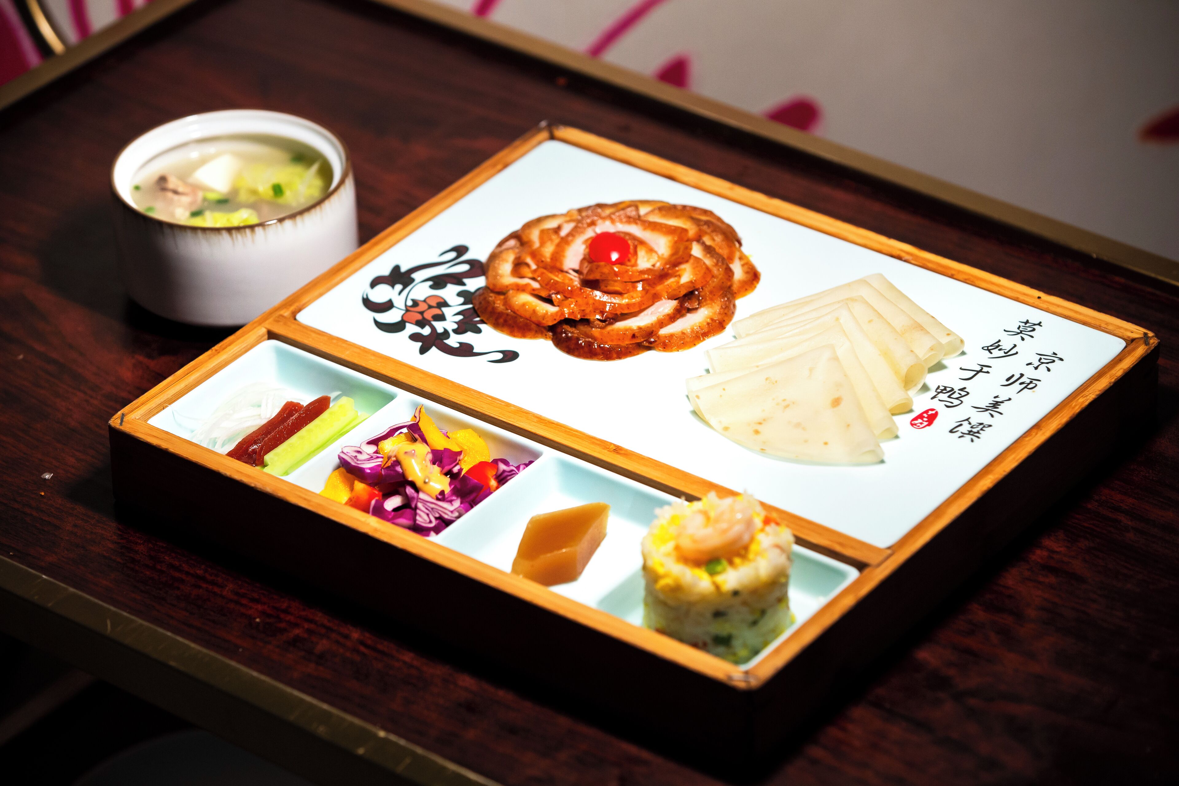 A traditional Japanese meal served on a wooden tray with a bowl of soup, a rolled omelette with a cherry, pickled vegetables, and a small dish of rice topped with a shrimp.