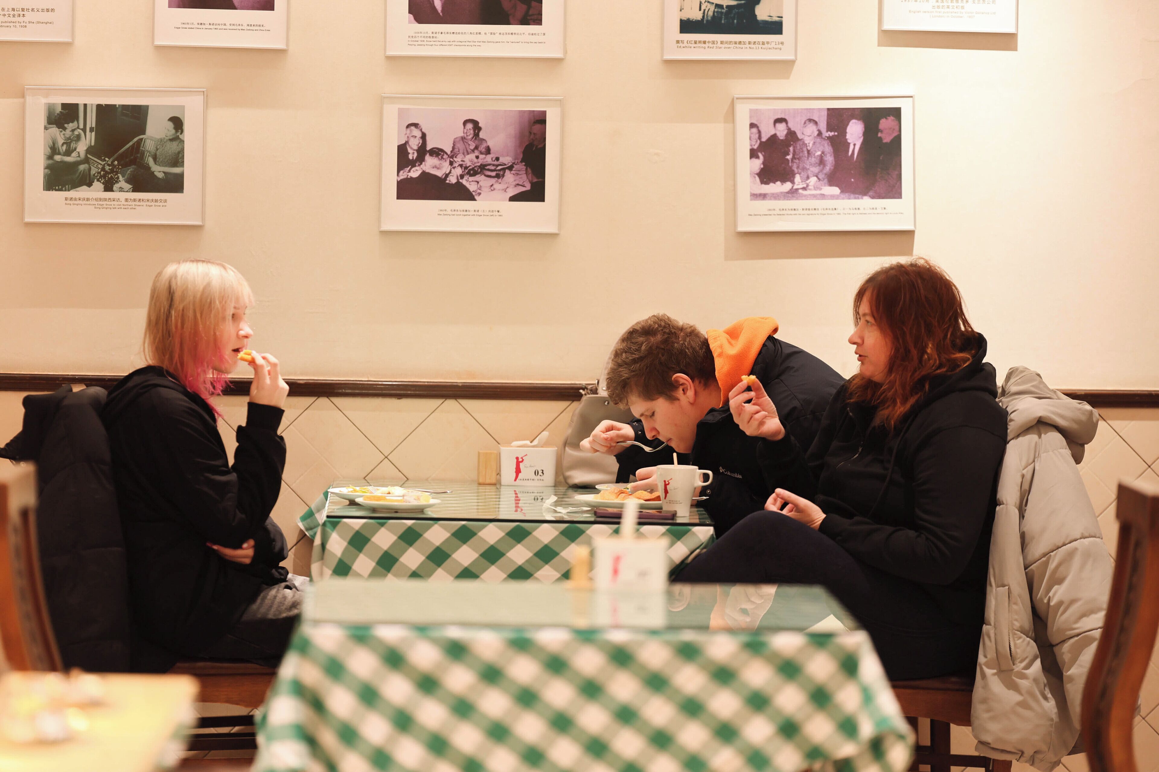 A dining area with checkered tablecloths, framed pictures on the wall, and a group of people seated at a table.