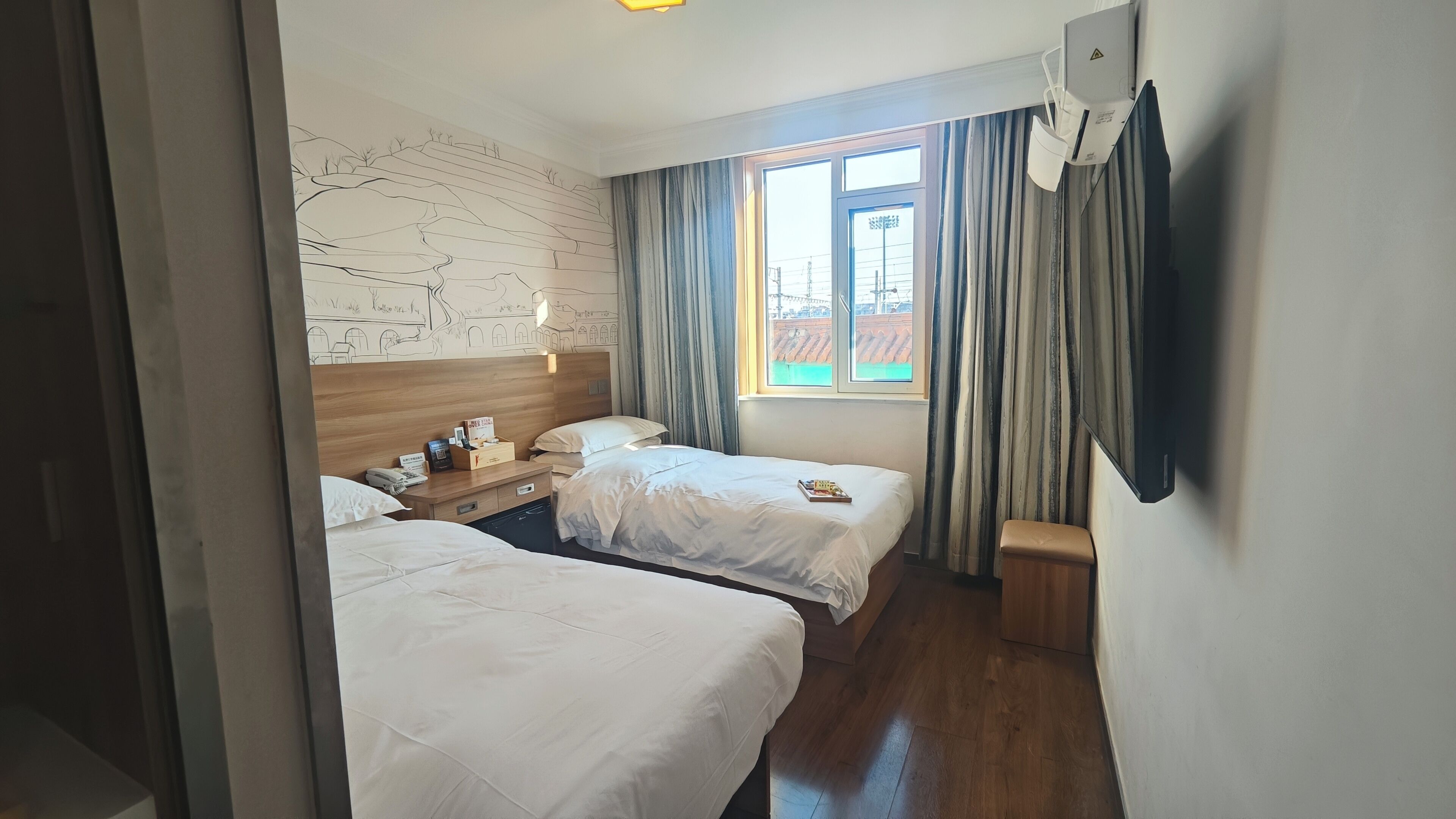 Een hotelkamer met twee bedden, een houten hoofdbord, een raam met gordijnen, een nachtkastje met een telefoon en een doos, en een aan de muur bevestigde televisie.