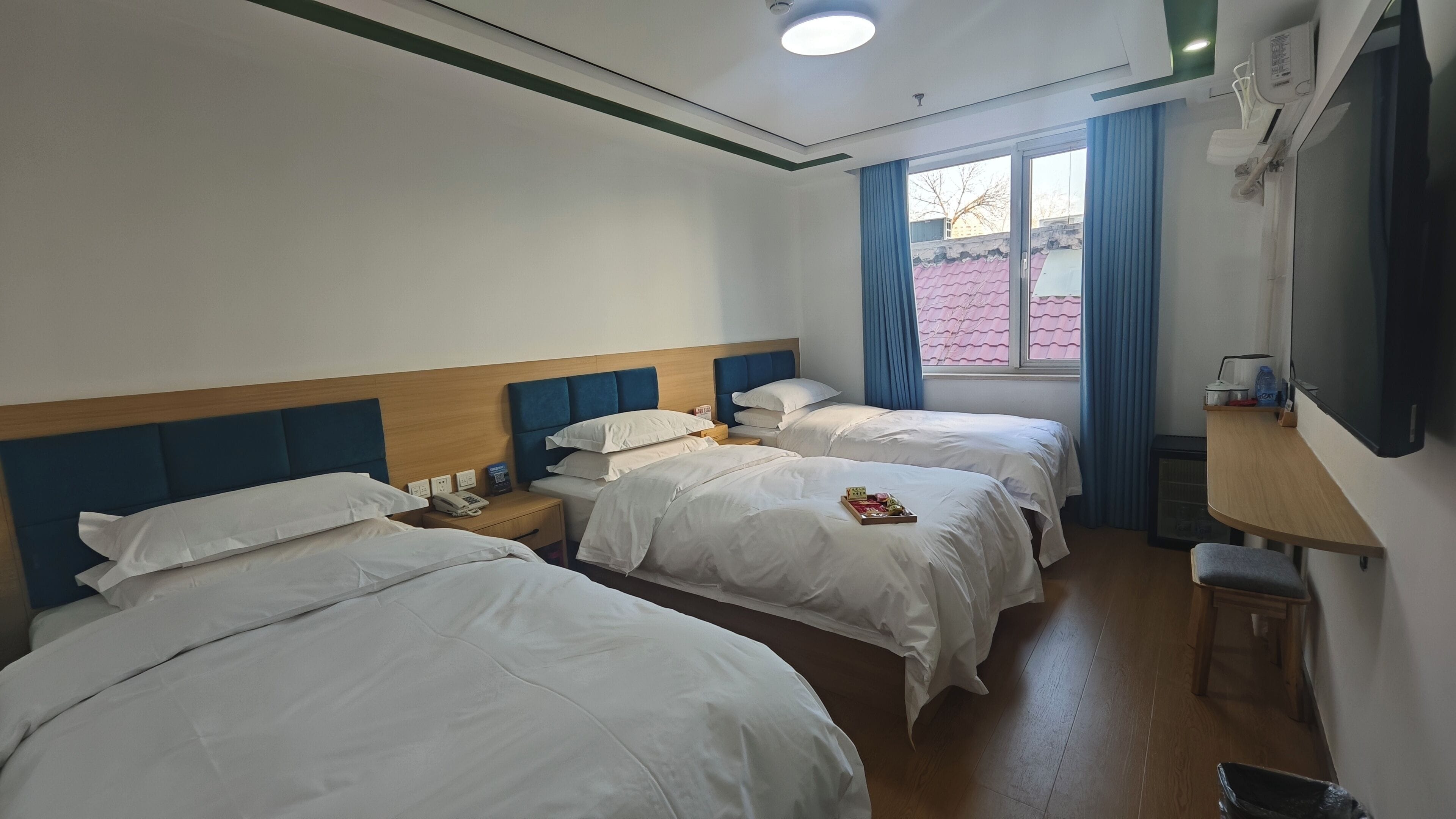 Ein Hotelzimmer mit drei Betten, einem Holzkopfstück, einem Schreibtisch mit Stuhl, einem Fenster mit Vorhängen und einem an der Wand befestigten Fernseher.