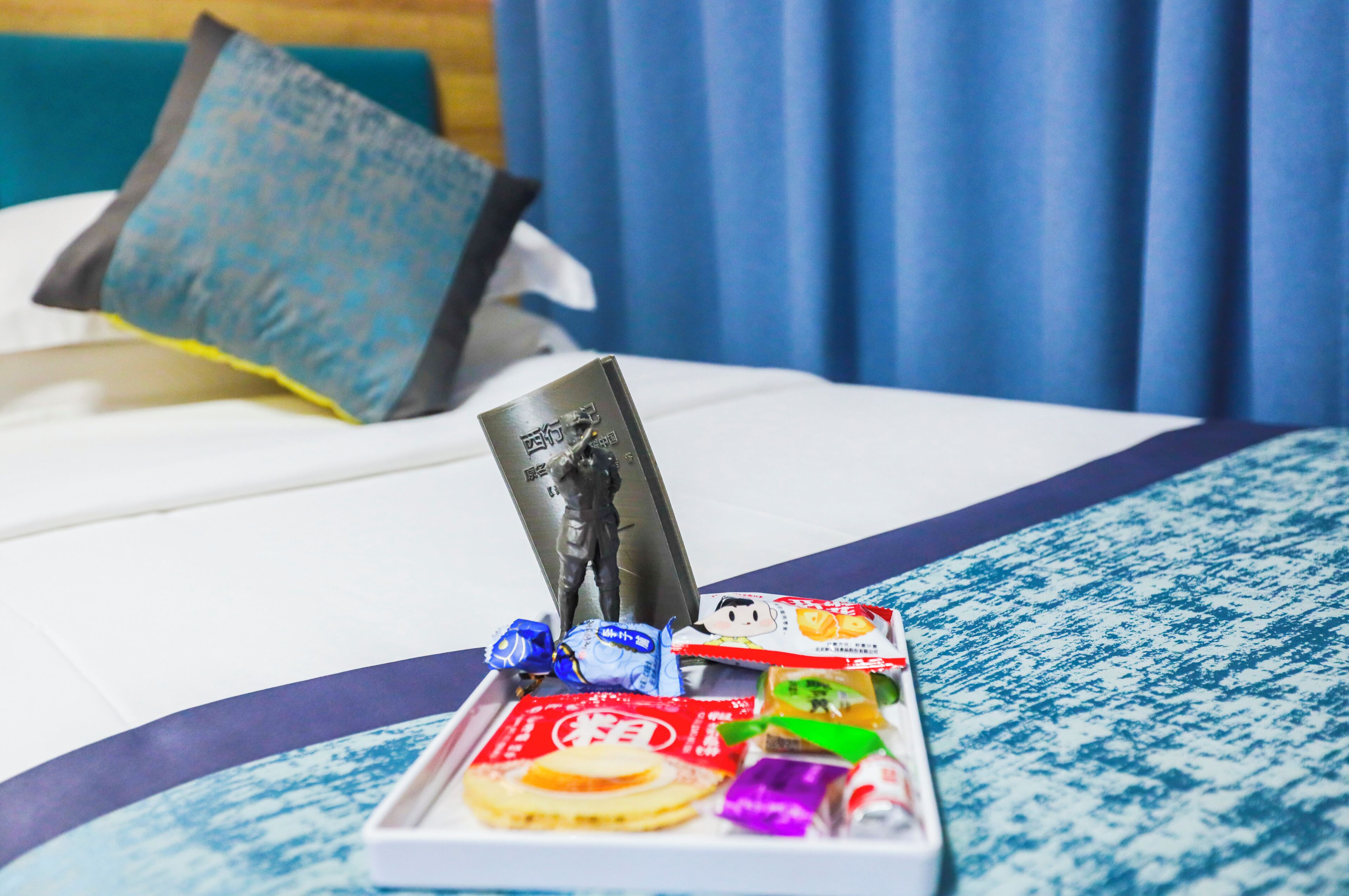 A neatly arranged tray of snacks and a small figurine on a bed with a blue and white patterned bedspread.