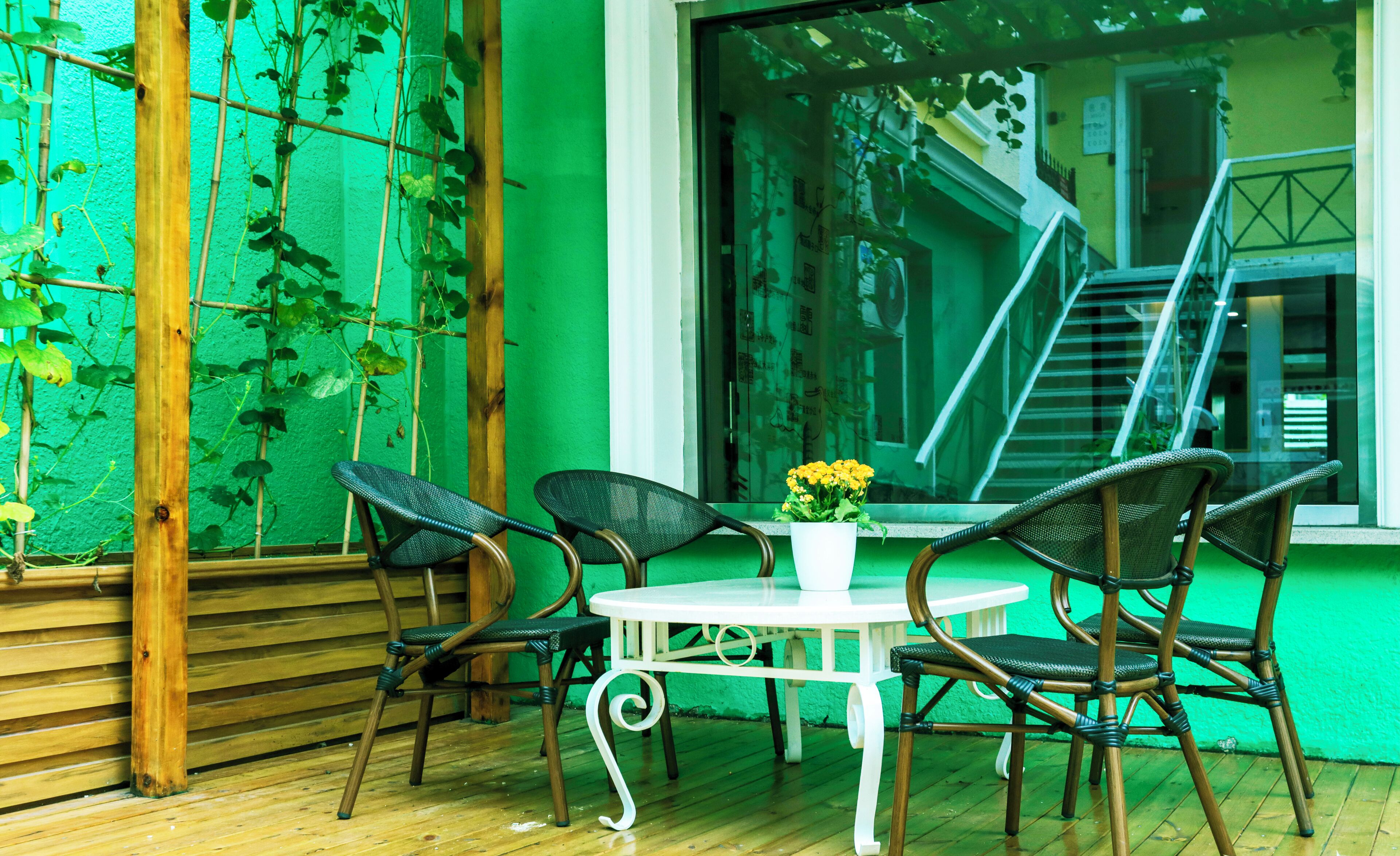 Una terraza de madera con sillas de mimbre y una mesa blanca, rodeada de paredes verdes y una ventana que da a una escalera.