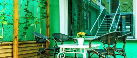 Una terraza de madera con sillas de mimbre y una mesa blanca, rodeada de paredes verdes y una ventana que da a una escalera.