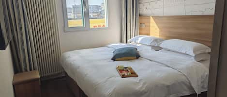 A hotel room with a bed, a wooden headboard, a window with curtains, and a wall with a drawing.
