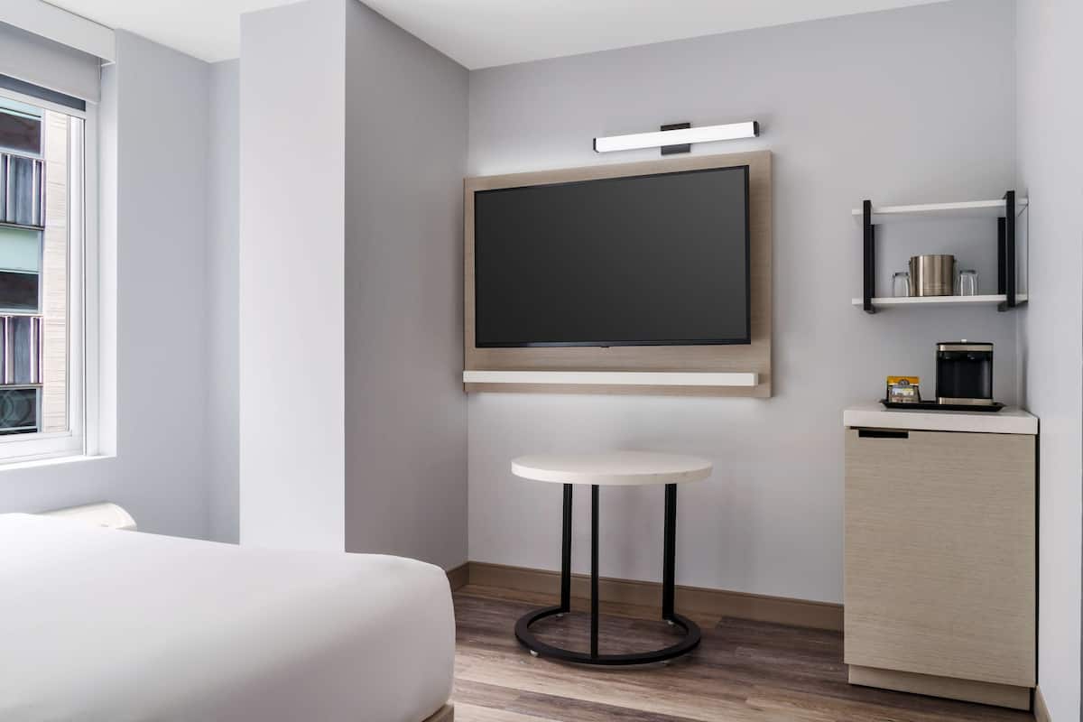 A modern hotel room with a bed, a small round table, a built-in TV, and a compact kitchenette.