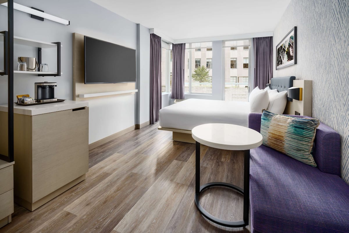 A modern hotel room with a bed, a sofa, a small table, a TV, and a window with curtains.