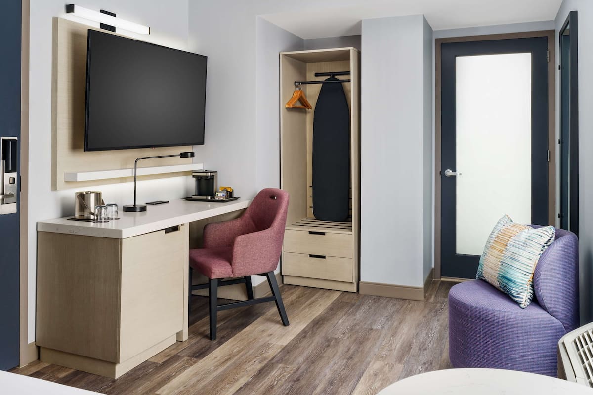 A modern room with a flat-screen TV mounted on the wall, a desk with a chair, a wardrobe with a hanging rack, and a purple sofa with a cushion.