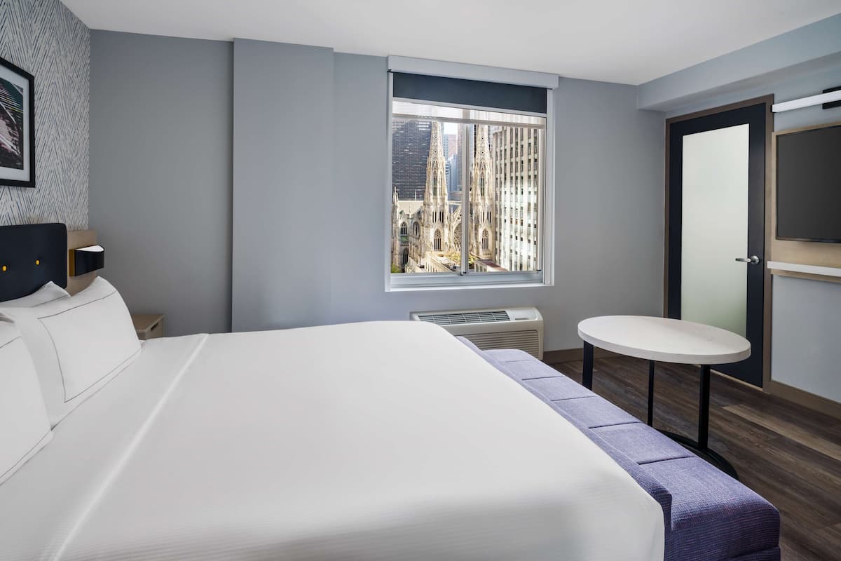 A modern hotel room with a large bed, a small round table, a window with a city view, and a mirror on the wall.