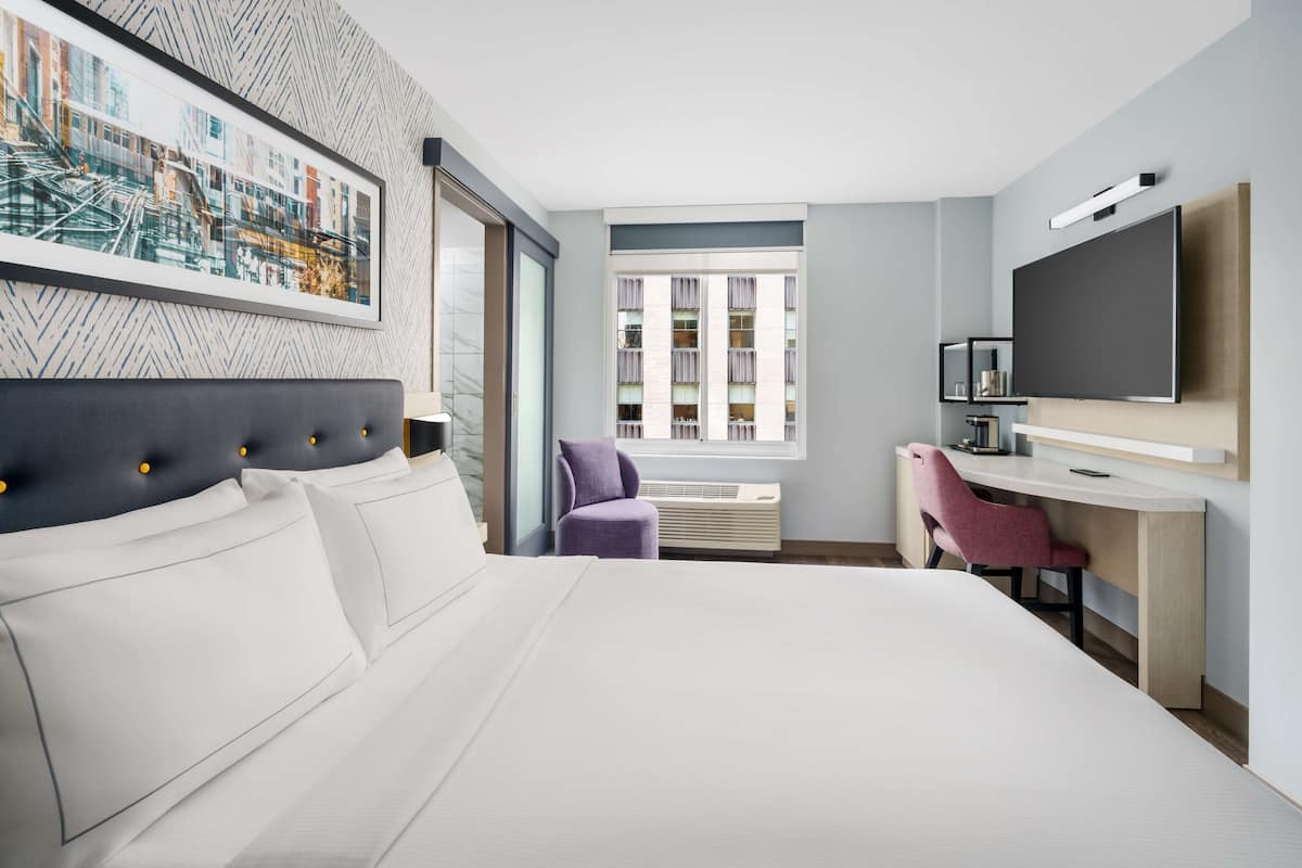 A modern hotel room with a large bed, a desk with a chair, a flat-screen TV, and a window with a view of buildings.