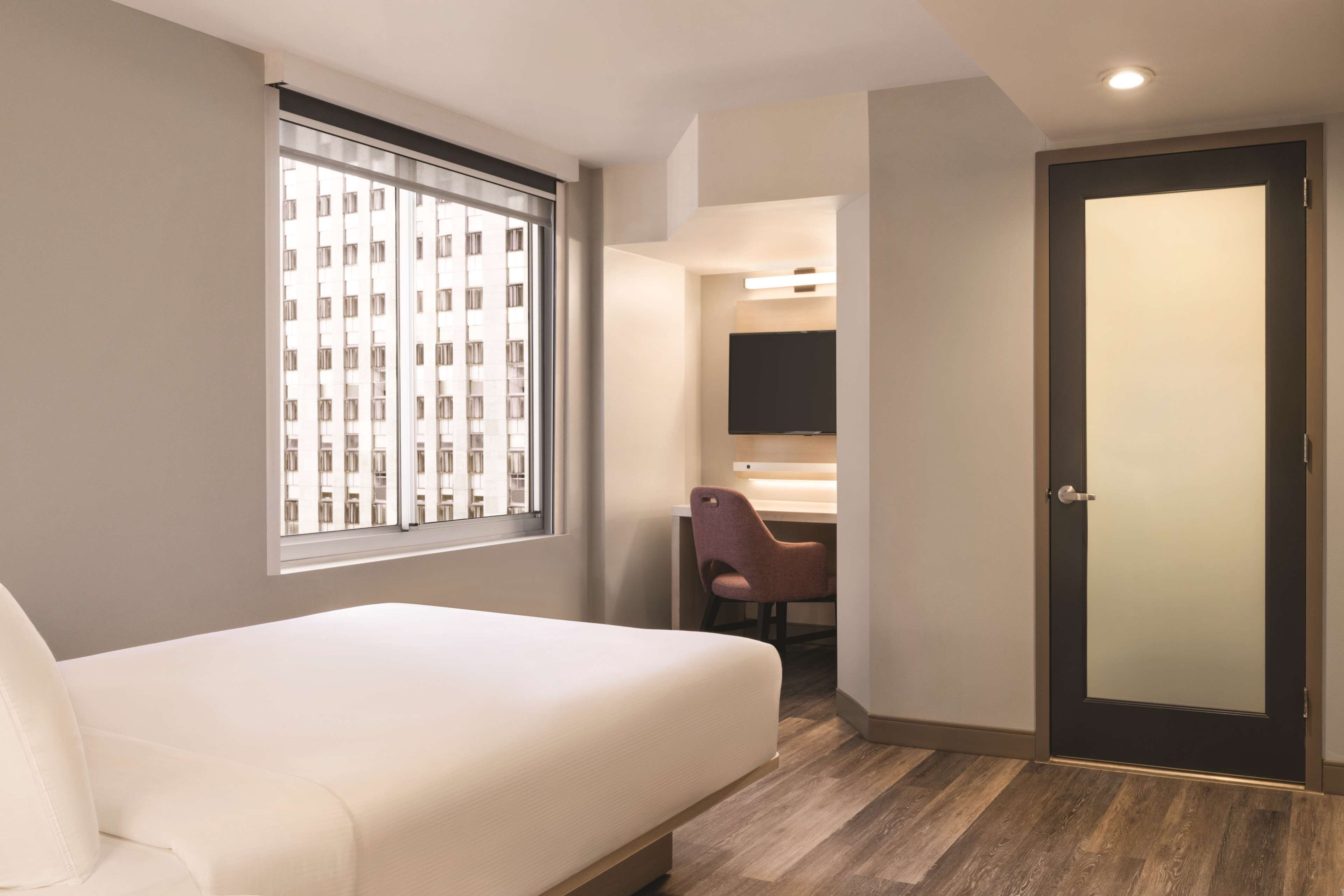 A modern hotel room with a bed, a desk, a chair, a TV, and a door.