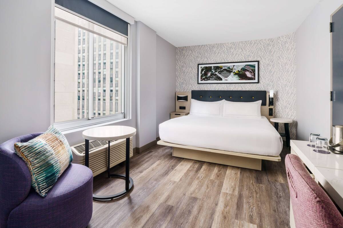 A modern hotel room with a large bed, a round table, a purple armchair, and a view of a building through the window.