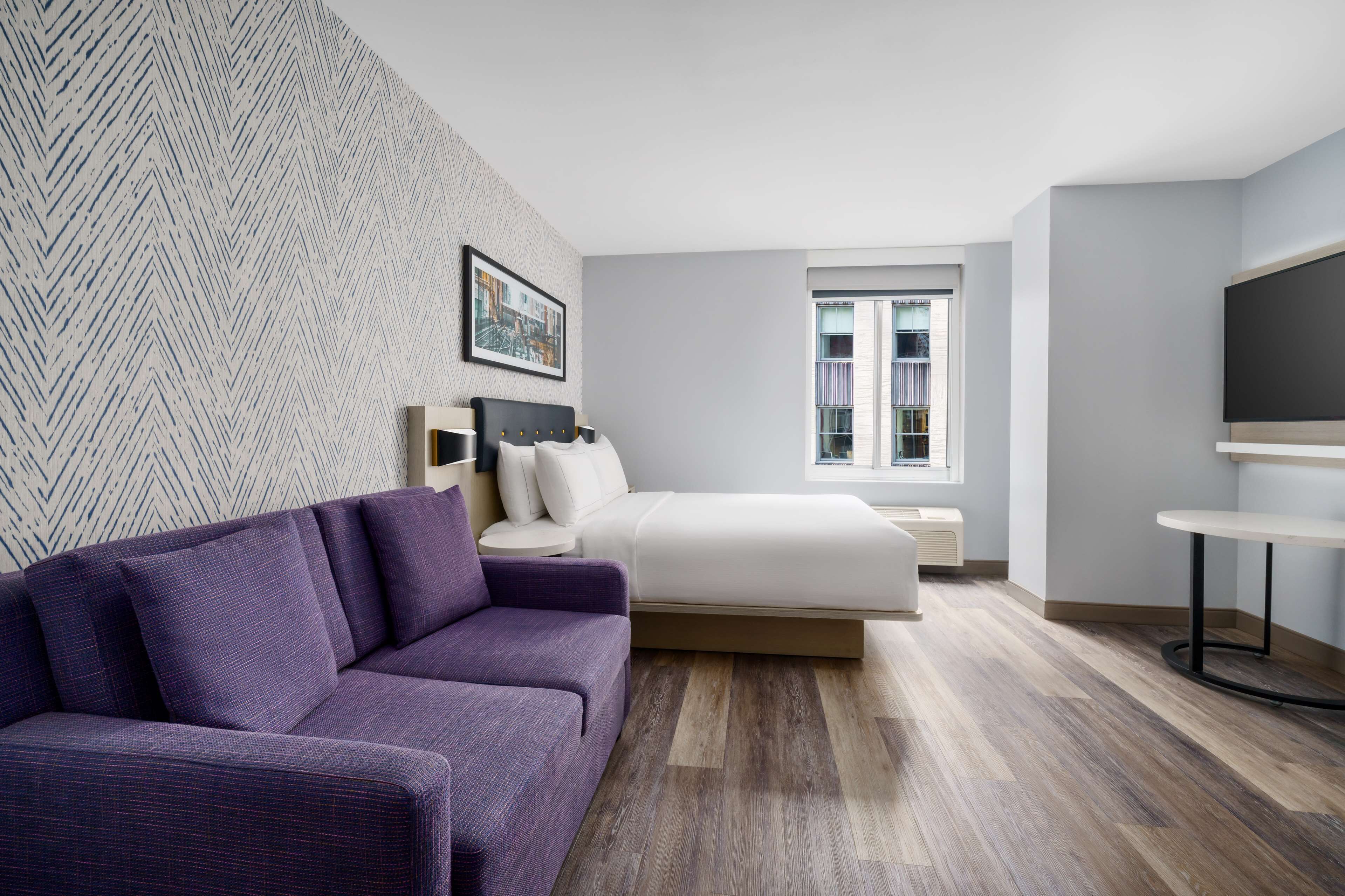 A modern hotel room with a purple sofa, a bed with a white headboard, a TV, and a window with a view of buildings.