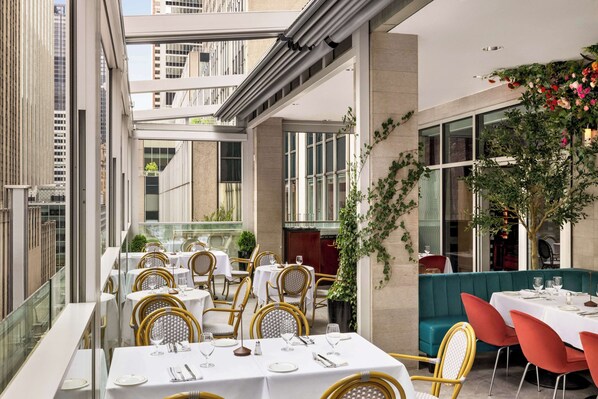 A restaurant with outdoor seating, white tables, wicker chairs, and a green cushioned bench. - DoubleTree by Hilton New York Midtown Fifth Ave (New York)