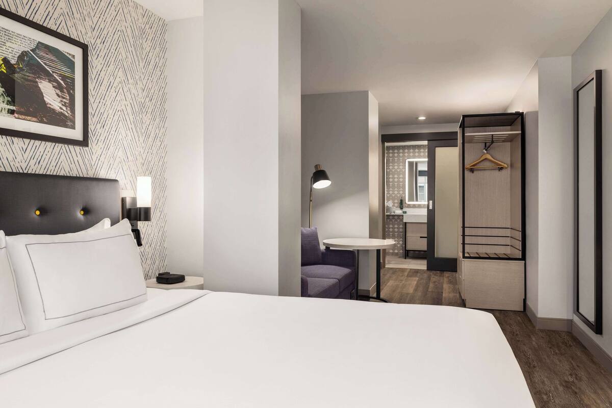 A modern hotel room with a bed, bedside table, lamp, and a view into the bathroom.