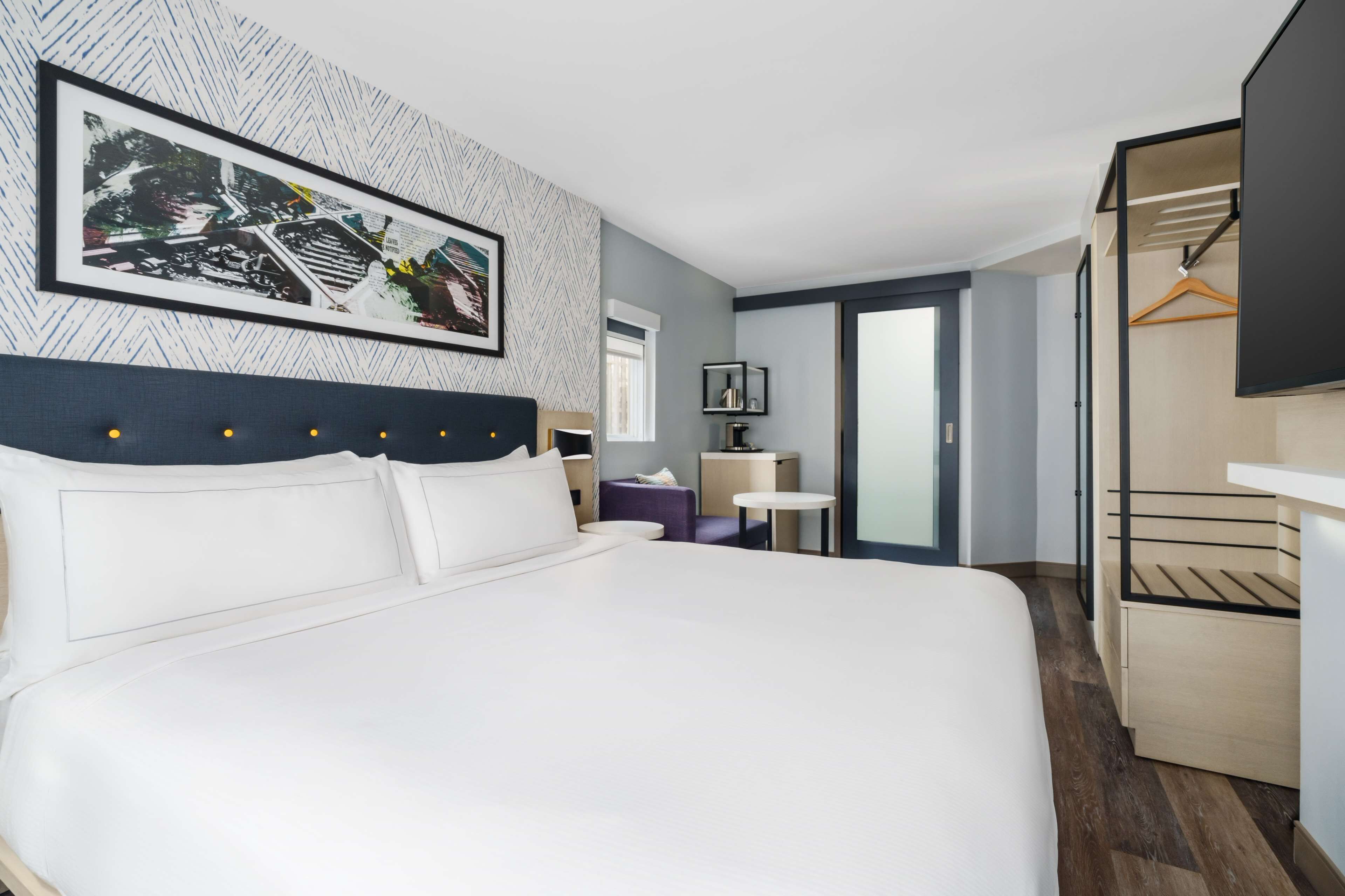 A modern hotel room with a large bed, a wardrobe, a small table, and a framed picture on the wall.