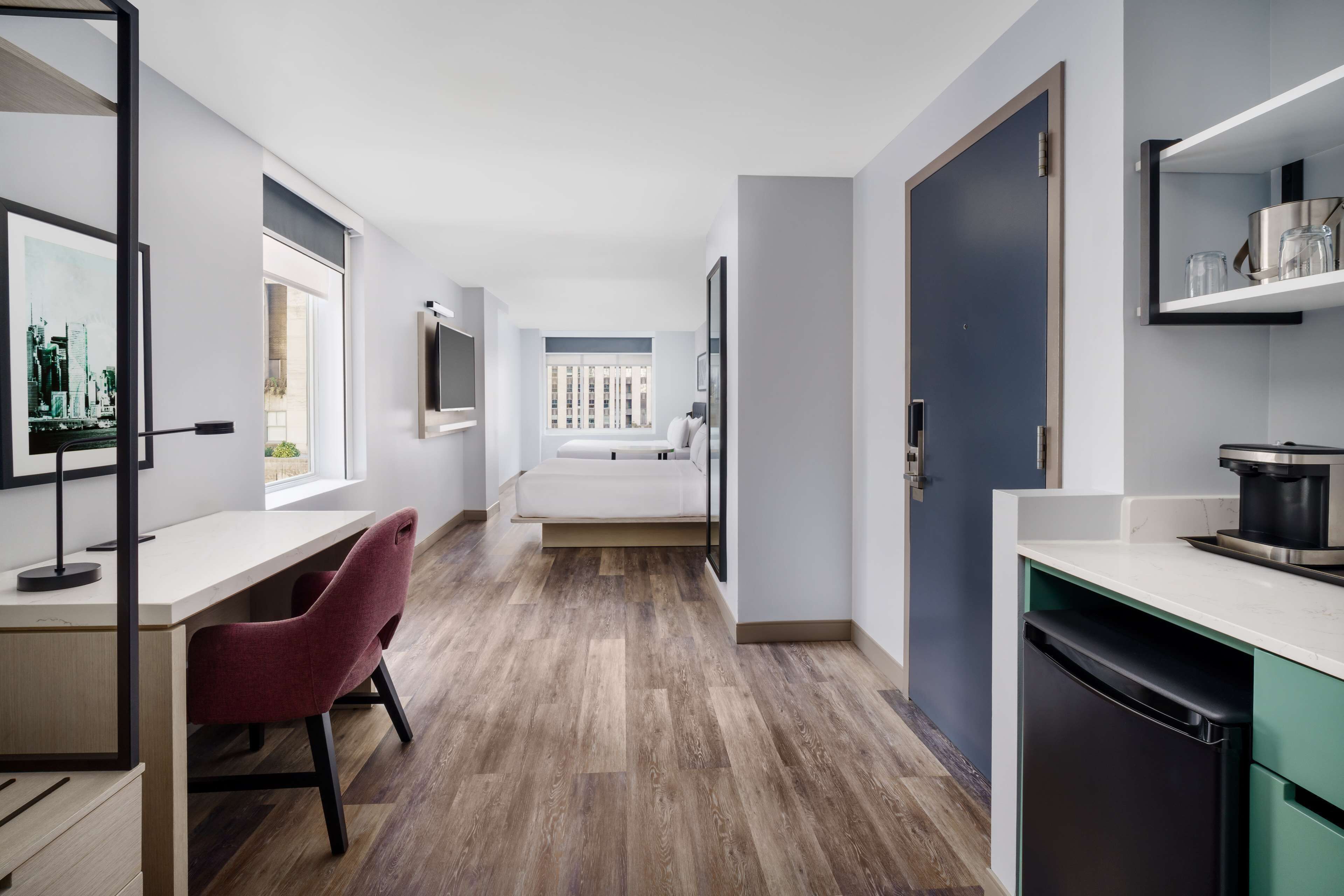 A modern apartment with a wooden floor, a desk with a chair, a bed, a kitchen area, and a view through a window.