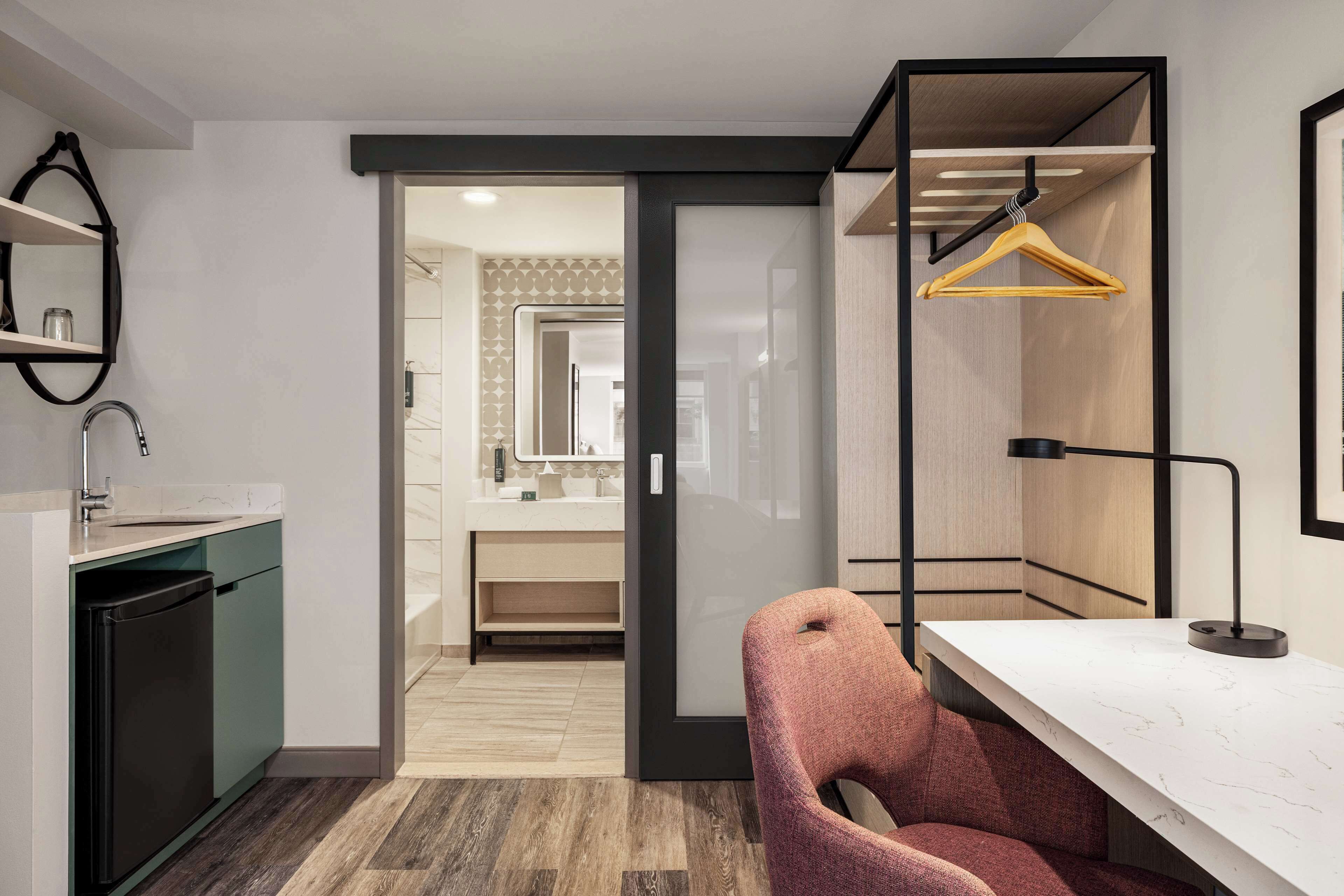 A modern hotel room with a kitchen area, a seating area with a chair, and a wardrobe with a hanging rack.