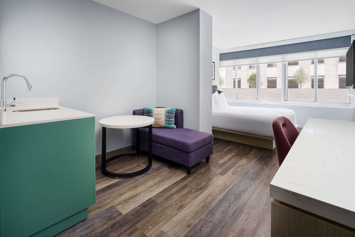 A modern hotel room with a kitchen area, a small round table with a purple ottoman, a white bed, and a large window offering a view of the outside.