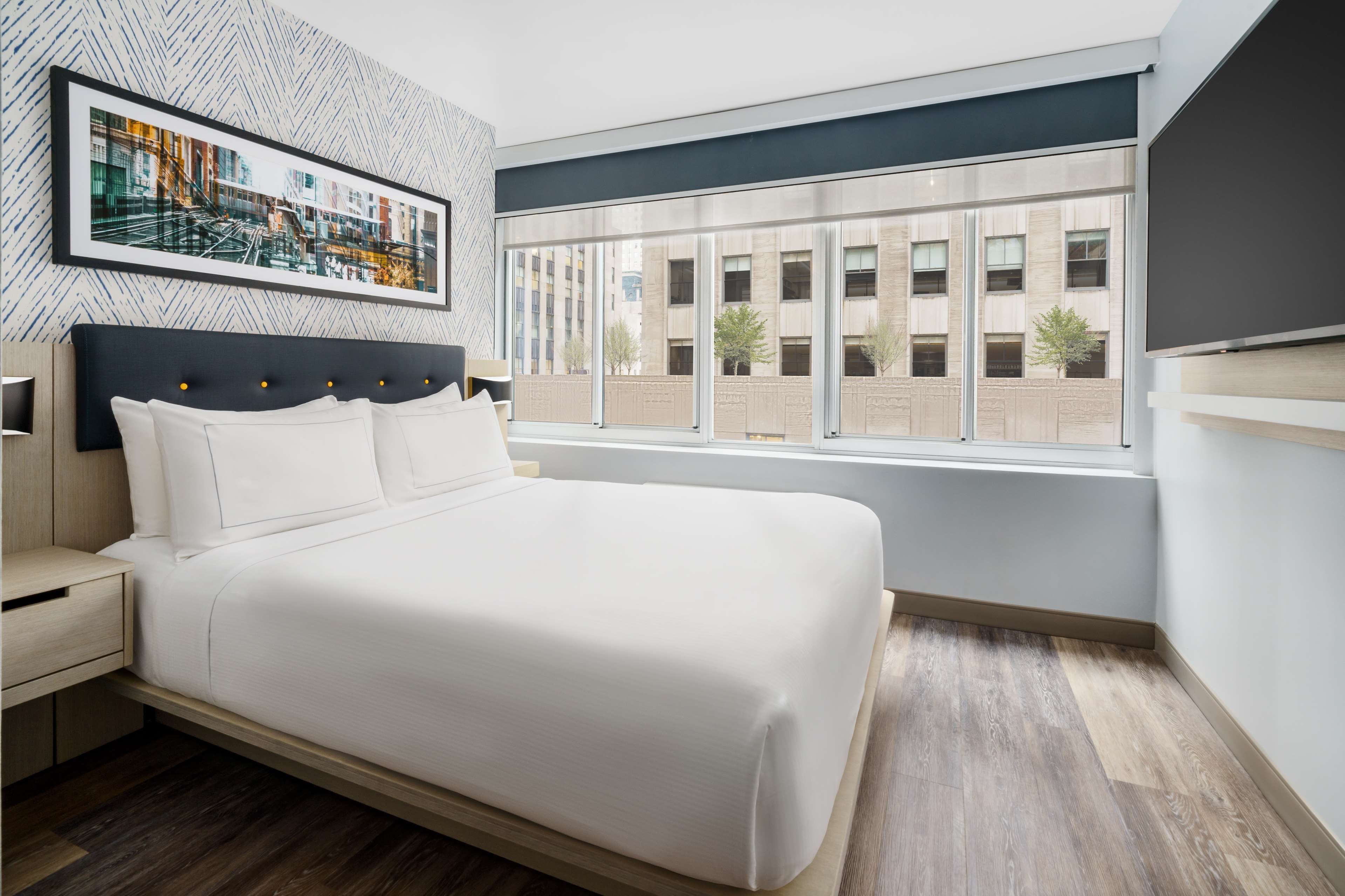 A modern bedroom with a large bed, a nightstand, a framed cityscape picture, and a window with a view of buildings.