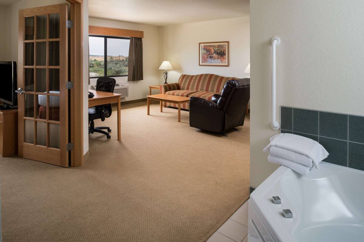 Habitación de hotel con puerta de madera, un sofá, un escritorio con silla, una lámpara y una bañera con toallas.