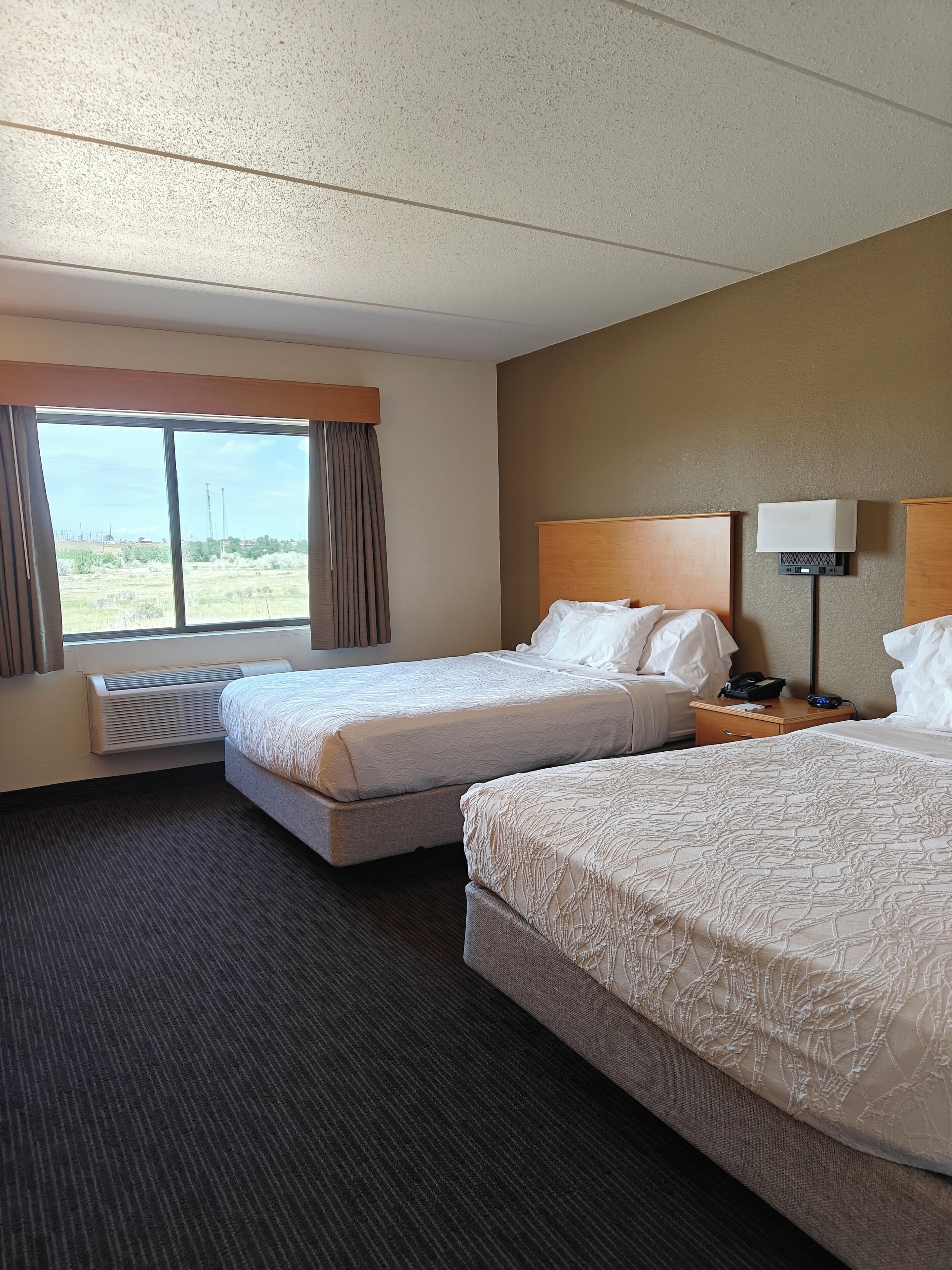A hotel room with two beds, a large window, a ceiling fan, and a bedside table with a lamp.