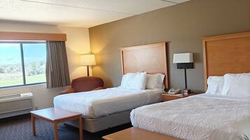 A hotel room with two beds, a desk, a chair, a lamp, and a view of the outdoors.