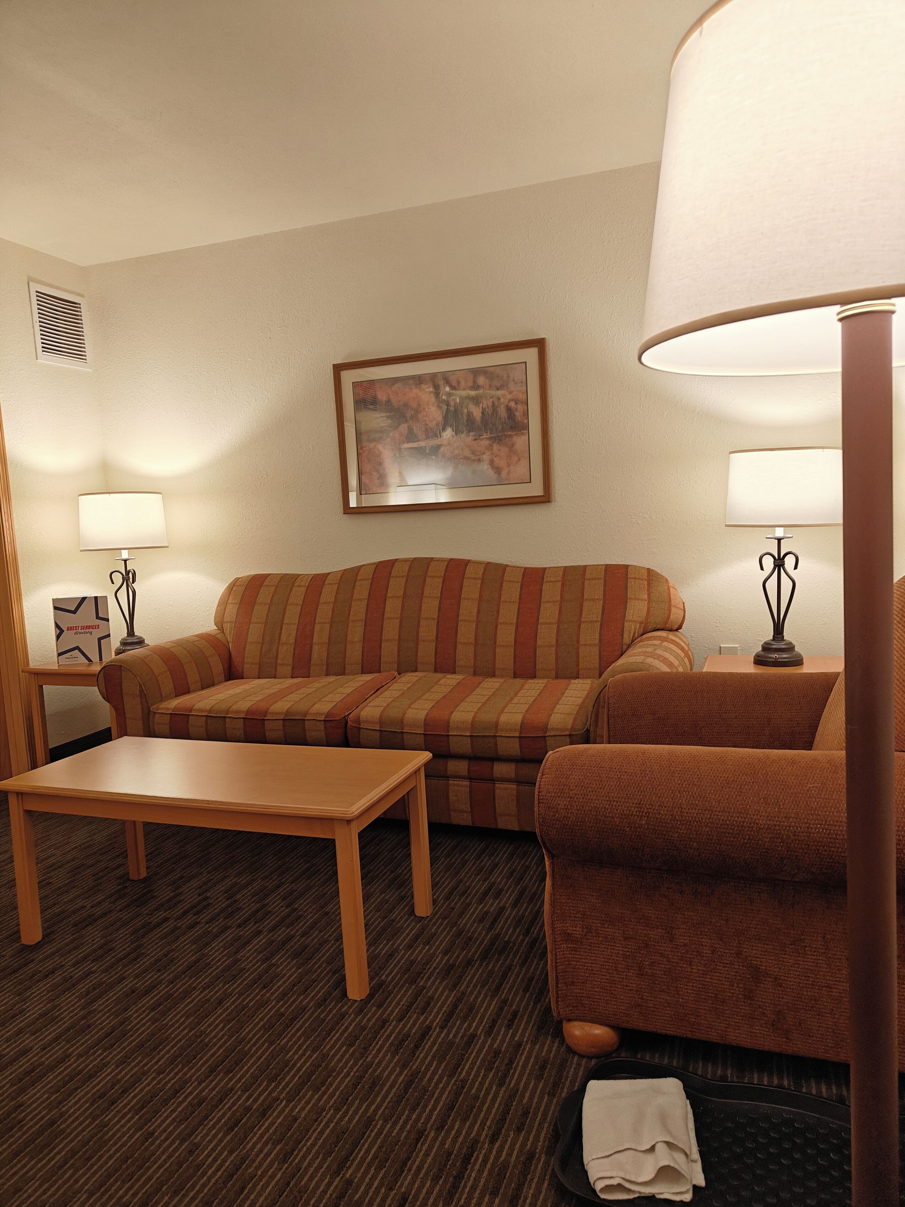Habitación de hotel con un sofá a rayas, una mesa de centro de madera, un cuadro en la pared y dos lámparas sobre mesitas de noche.