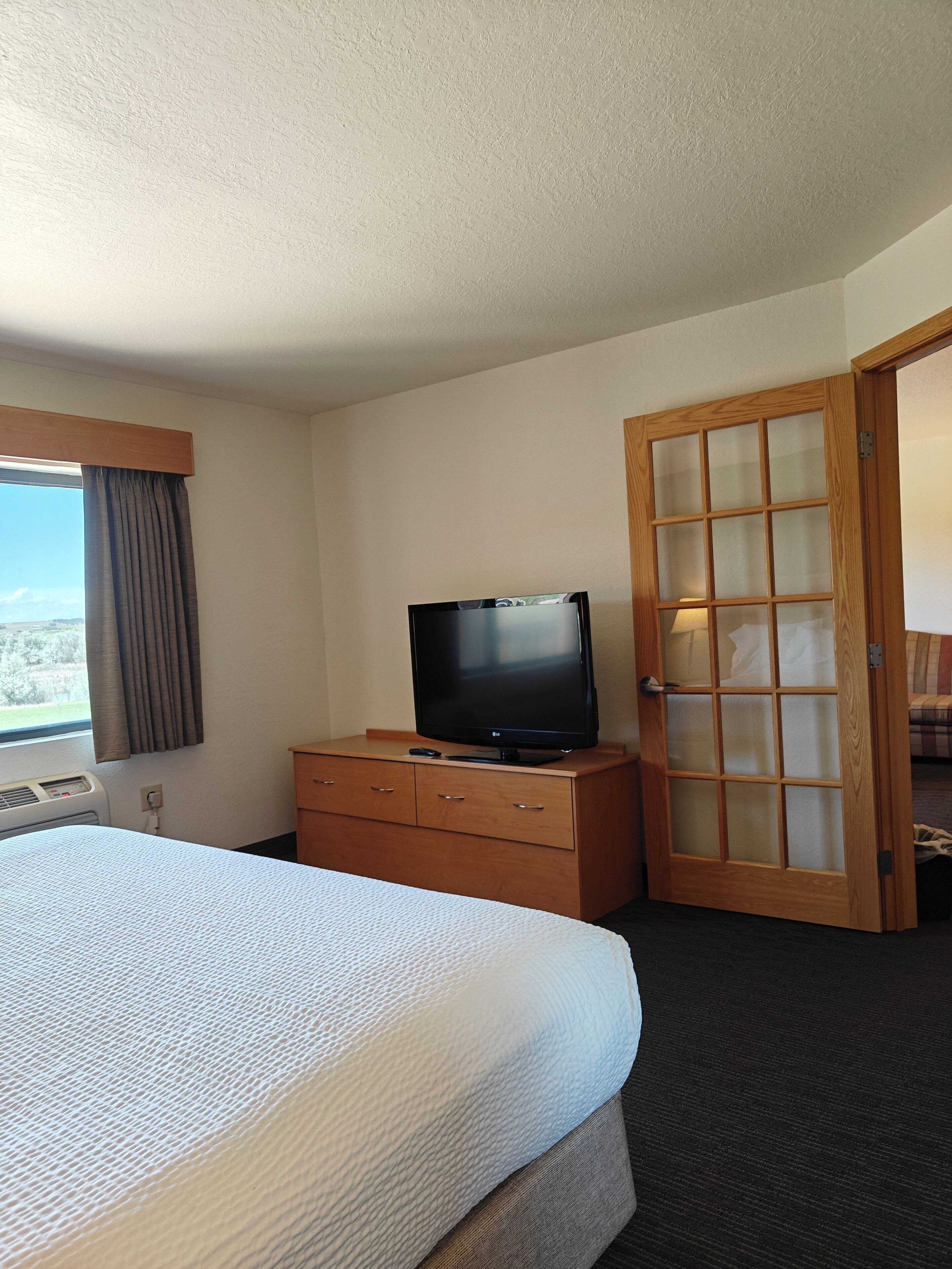Ein Hotelzimmer mit einem Bett, einem Fernseher auf einer Holzkommode, einem Fenster mit Vorhängen und einer Tür mit satinierten Glasscheiben.