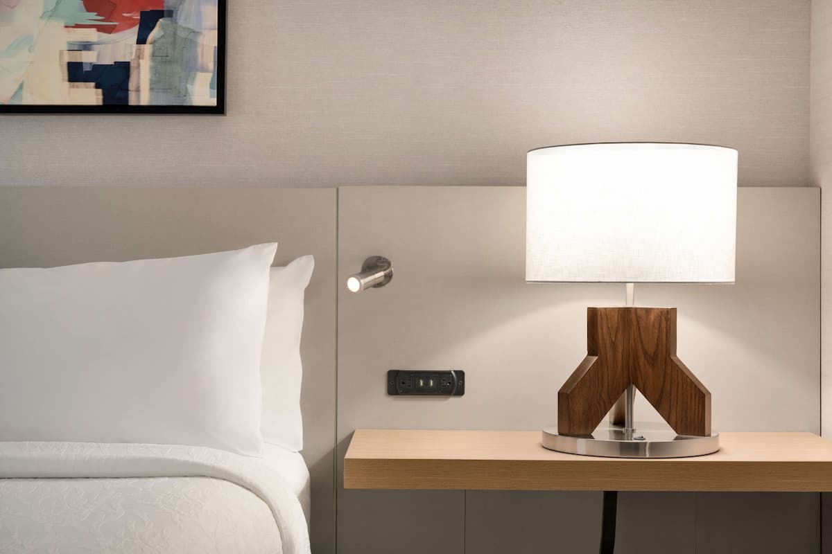 A neatly made bed with white linens, a bedside table with a modern lamp, and a power outlet on the wall.