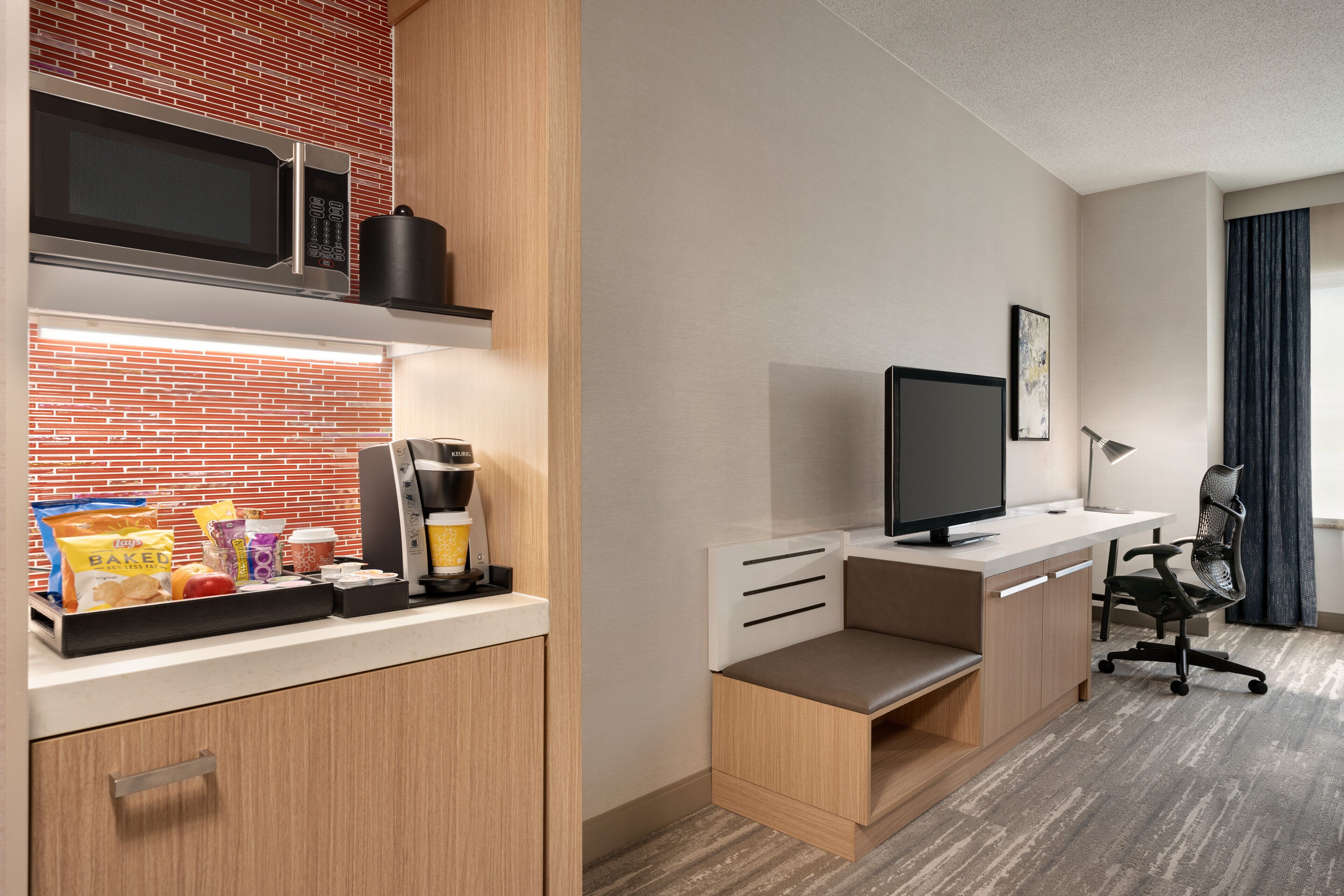 A compact hotel room with a kitchenette, a desk with a computer, and a window with curtains.