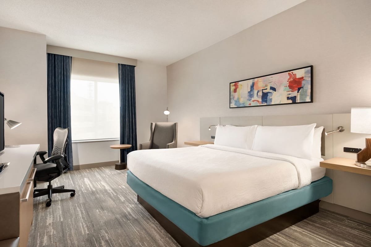 A hotel room with a bed, desk, chair, and artwork on the wall.