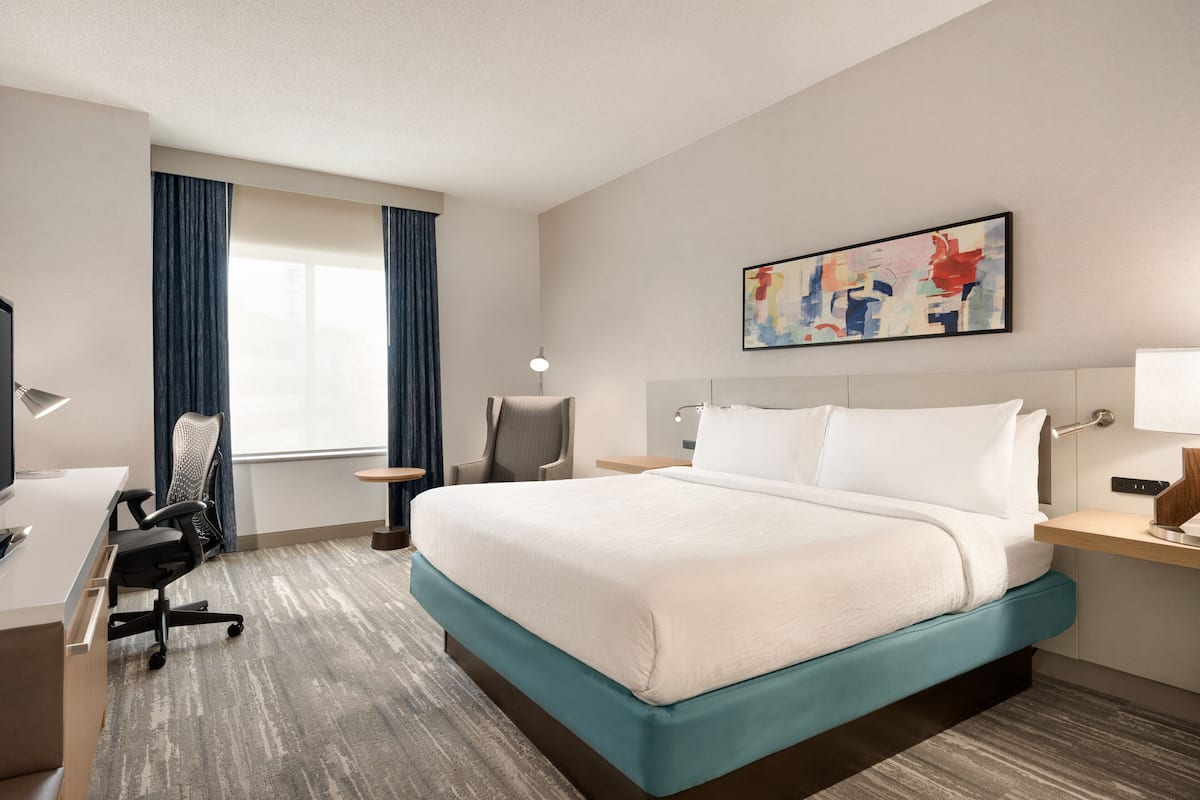 A hotel room with a bed, desk, chair, and a painting on the wall.