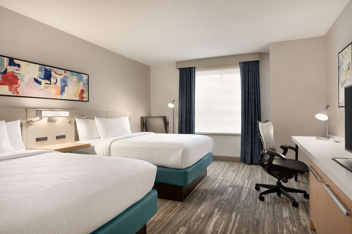 A hotel room with two beds, a desk, a chair, a lamp, and a large abstract painting on the wall.