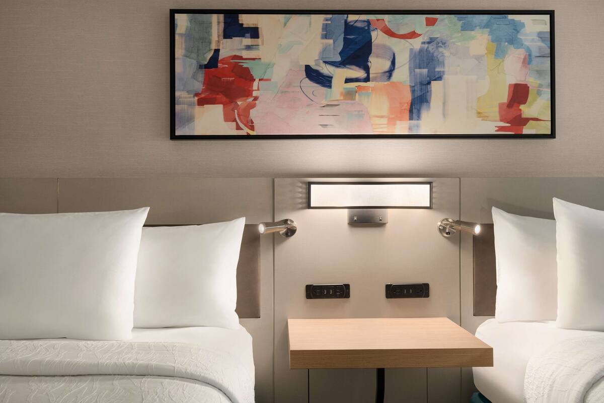 A hotel room with a bed, bedside tables, a small desk, and a colorful abstract painting on the wall.