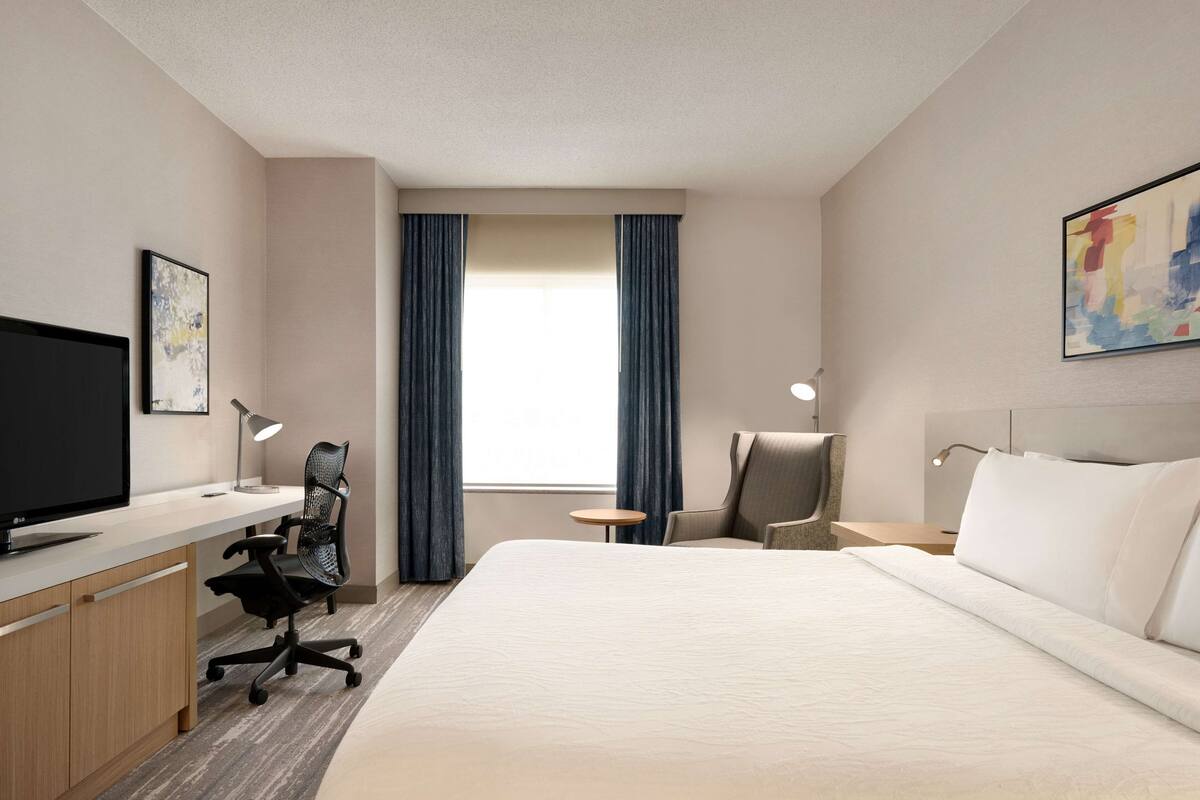 A hotel room with a bed, desk, chair, TV, and artwork on the walls.