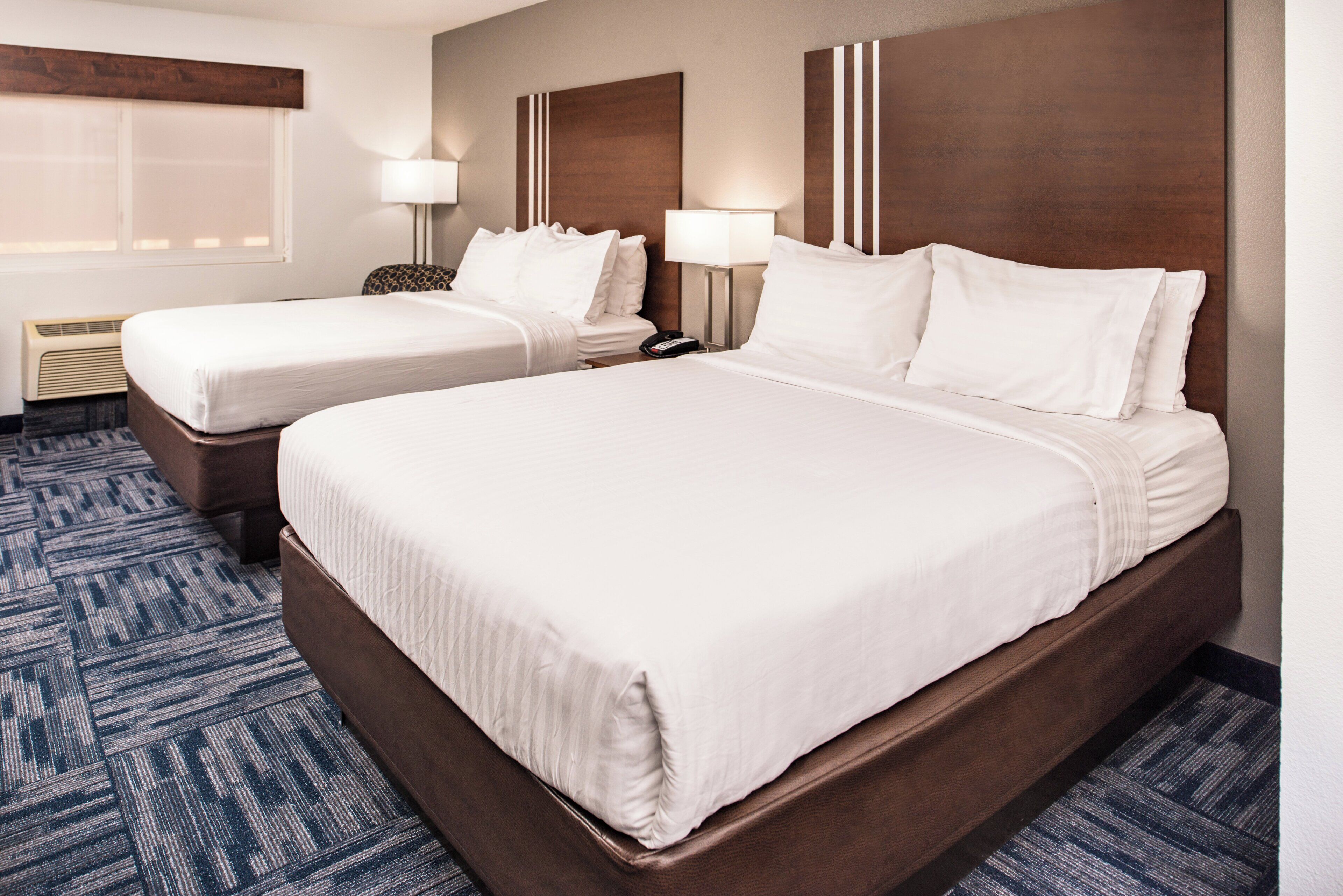 A hotel room with two beds, a nightstand, a lamp, and a window with blinds.