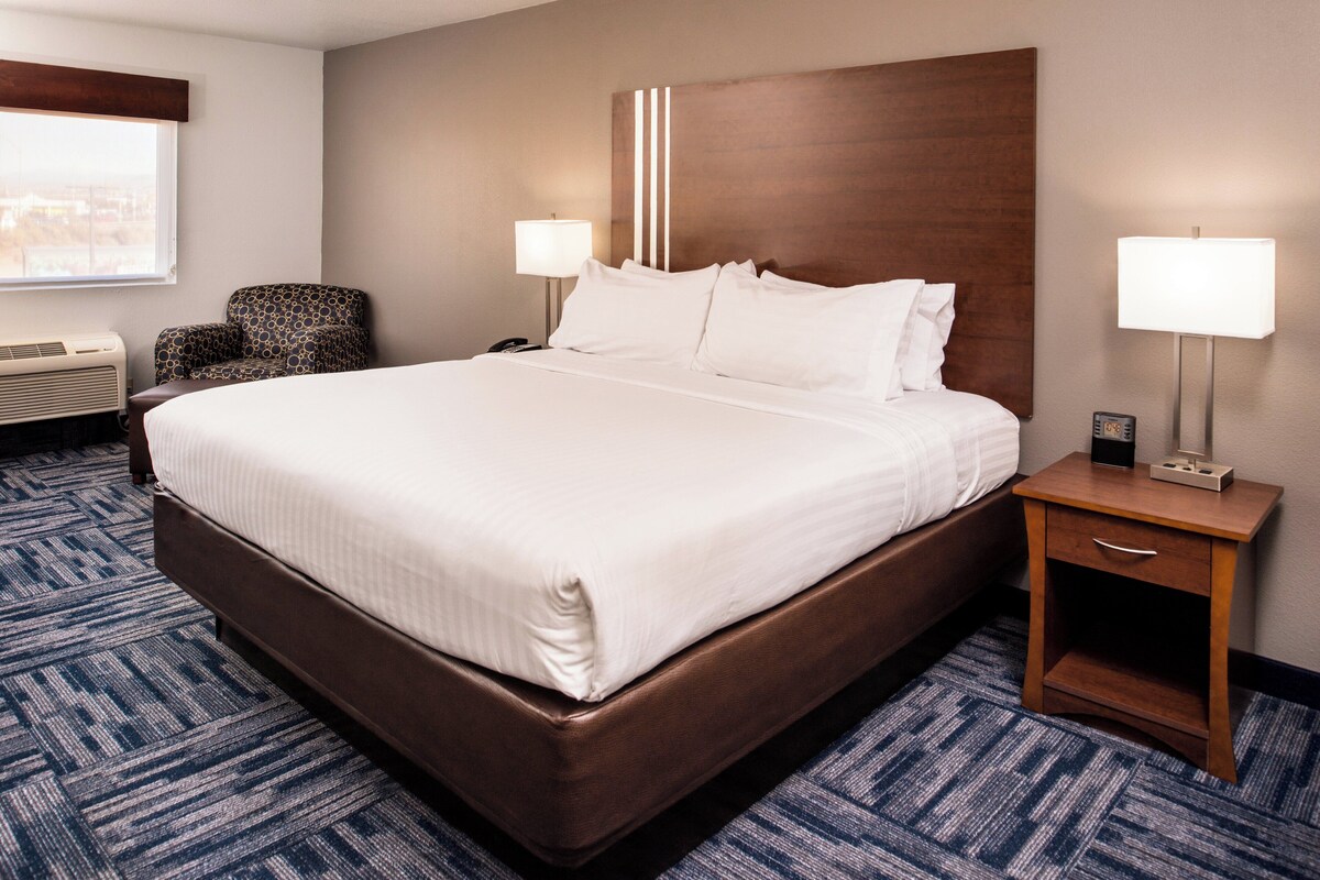 Una habitación de hotel con una cama grande, una silla, una mesita de noche, una lámpara y una ventana.