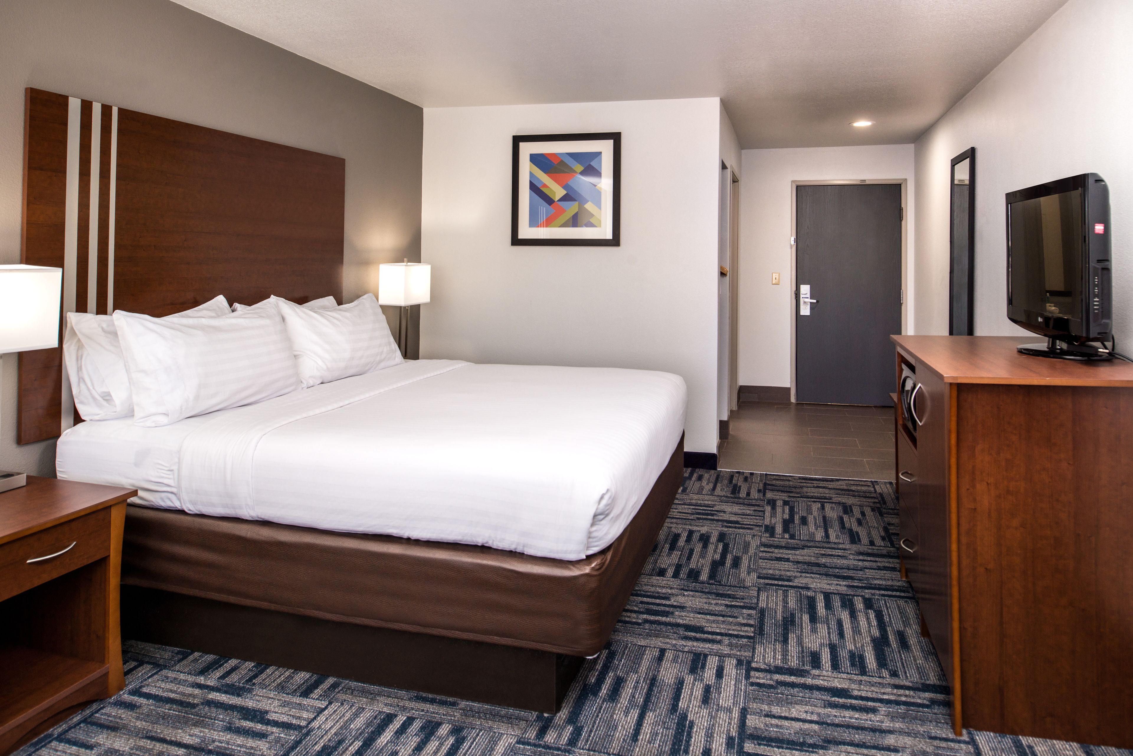 A hotel room with a large bed, a wooden headboard, a desk with a lamp, a television, and a framed abstract painting on the wall.