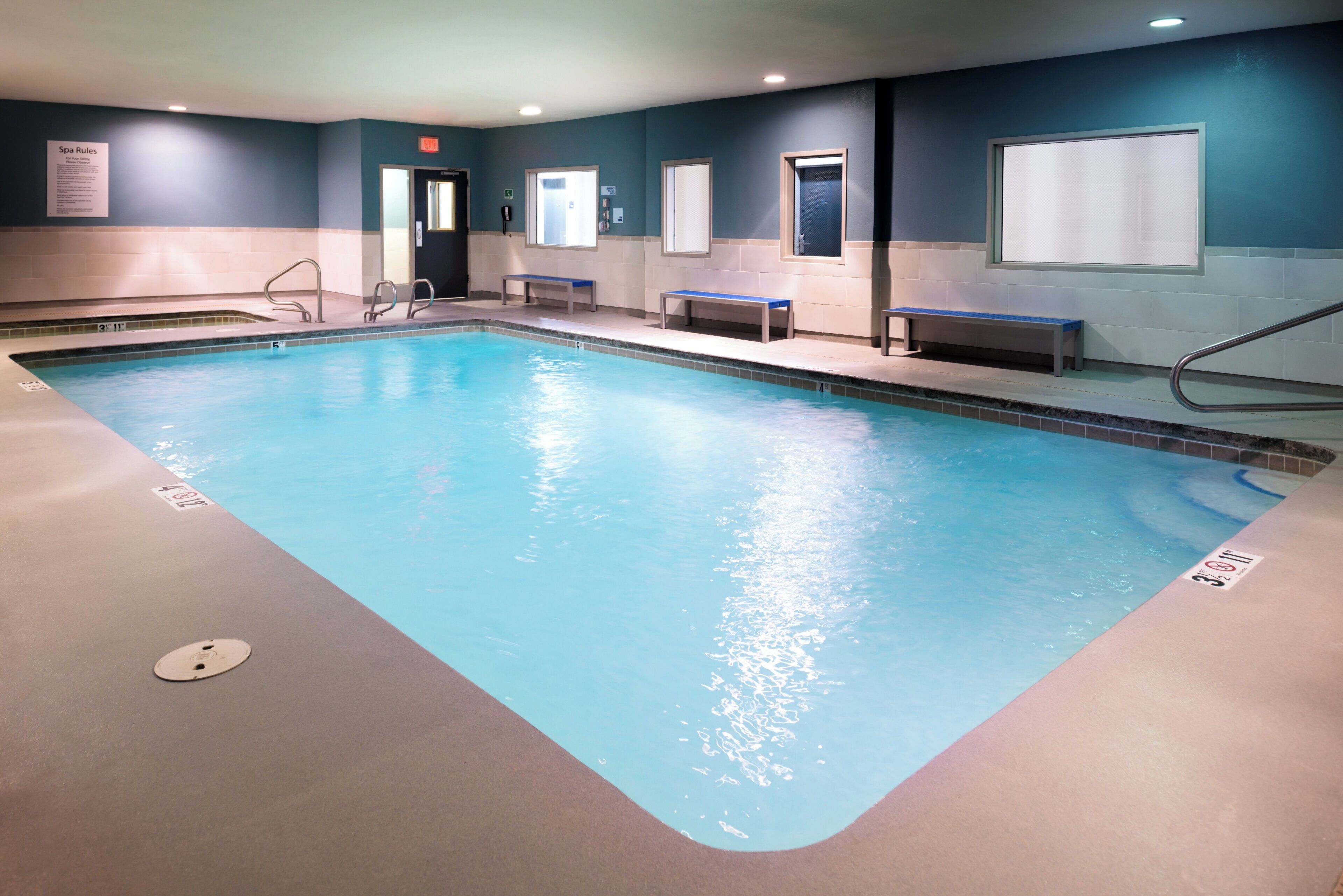 A large indoor swimming pool with clear blue water, surrounded by walls with windows and benches.