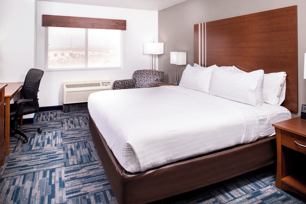 A hotel room with a large bed, a desk, a chair, a lamp, and a view of the city through the window.
