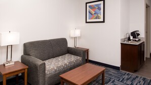 A hotel room with a gray sofa, two bedside tables with lamps, a wooden coffee table, a patterned rug, and a small kitchenette with a coffee maker. - Holiday Inn Express Hotel & Suites Alamogordo Hwy 54/70 by IHG (Alamogordo)