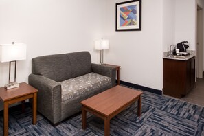 Habitación de hotel con un sofá gris, dos mesitas de noche con lámparas, una mesa de centro de madera, una alfombra estampada y una pequeña kitchenette con cafetera.