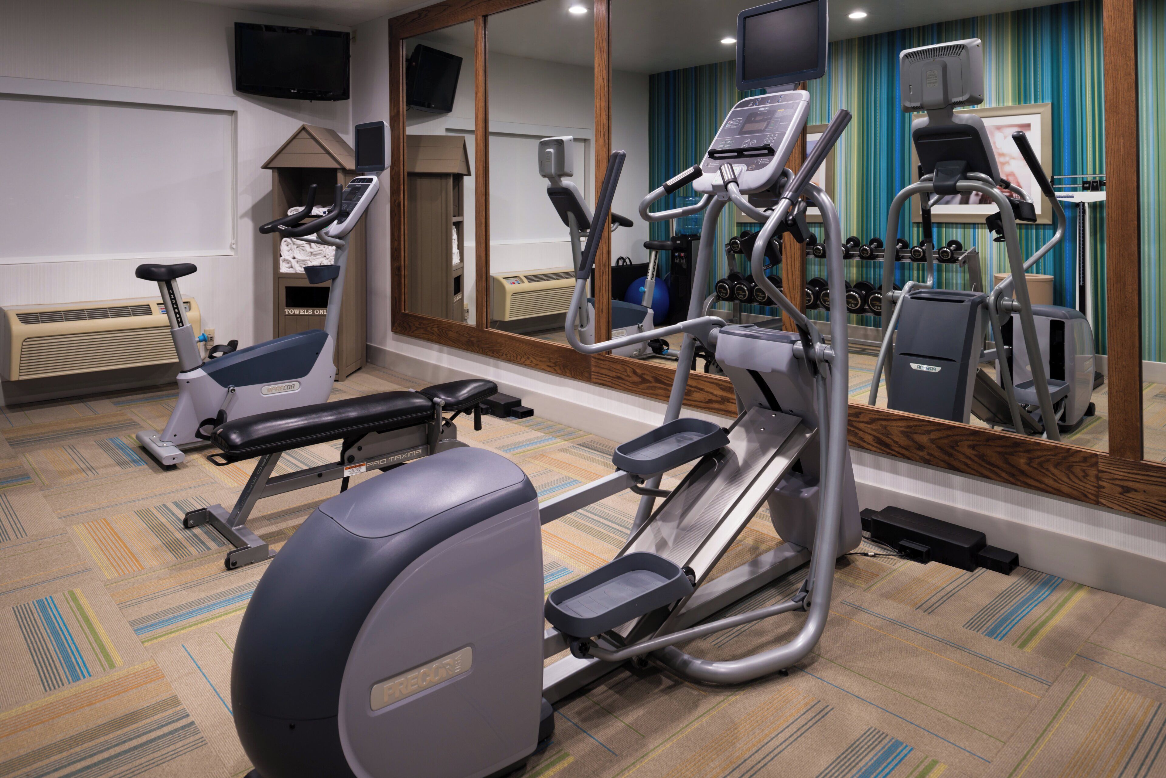 A gym with cardio machines, a mirror, and a wall-mounted TV.