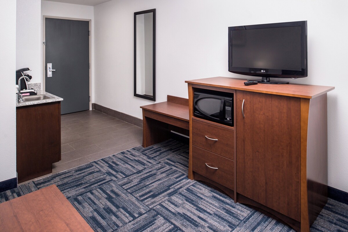 A hotel room with a wooden desk, a microwave, a television, a sink, and a mirror.