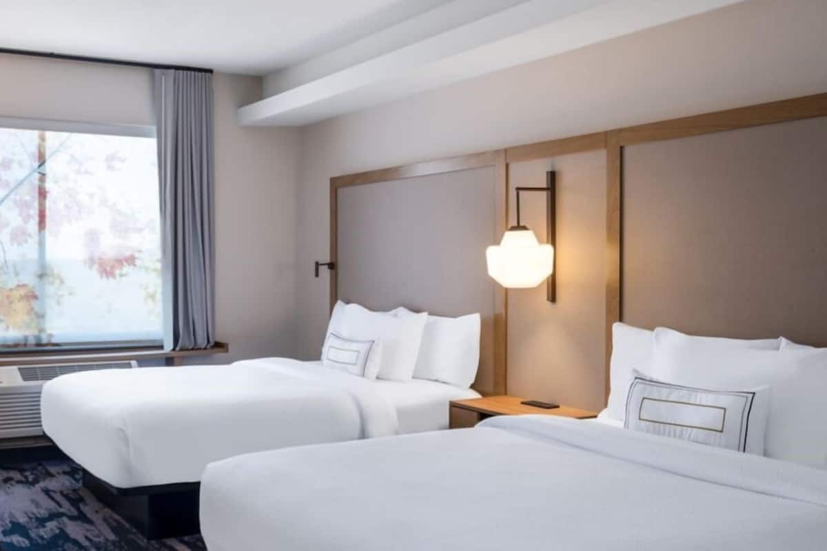 A hotel room with two beds, a large window with curtains, a wall-mounted light fixture, and a wooden headboard.