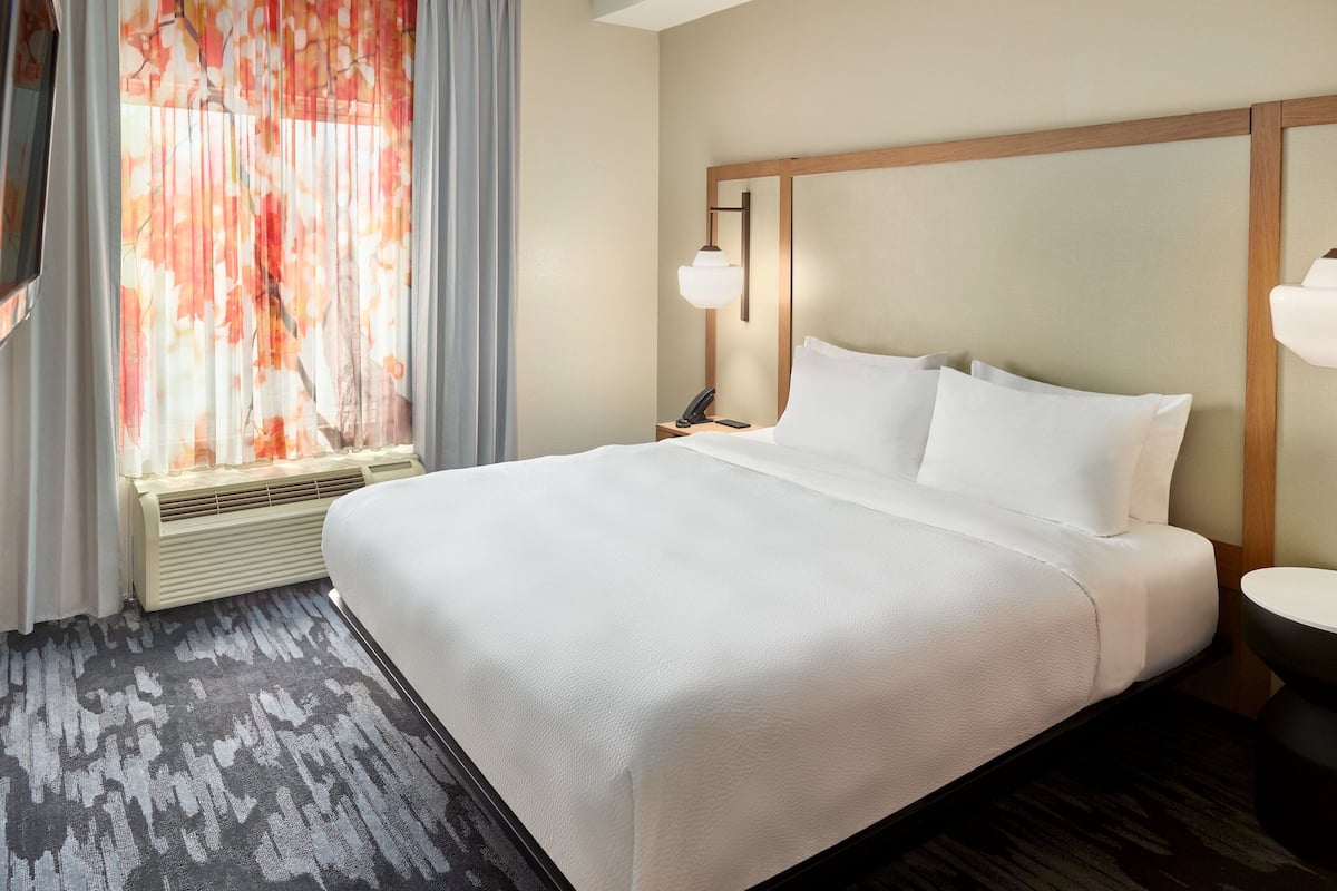 A hotel room with a large bed, a bedside table with a lamp, a window with curtains, and a wall-mounted air conditioning unit.
